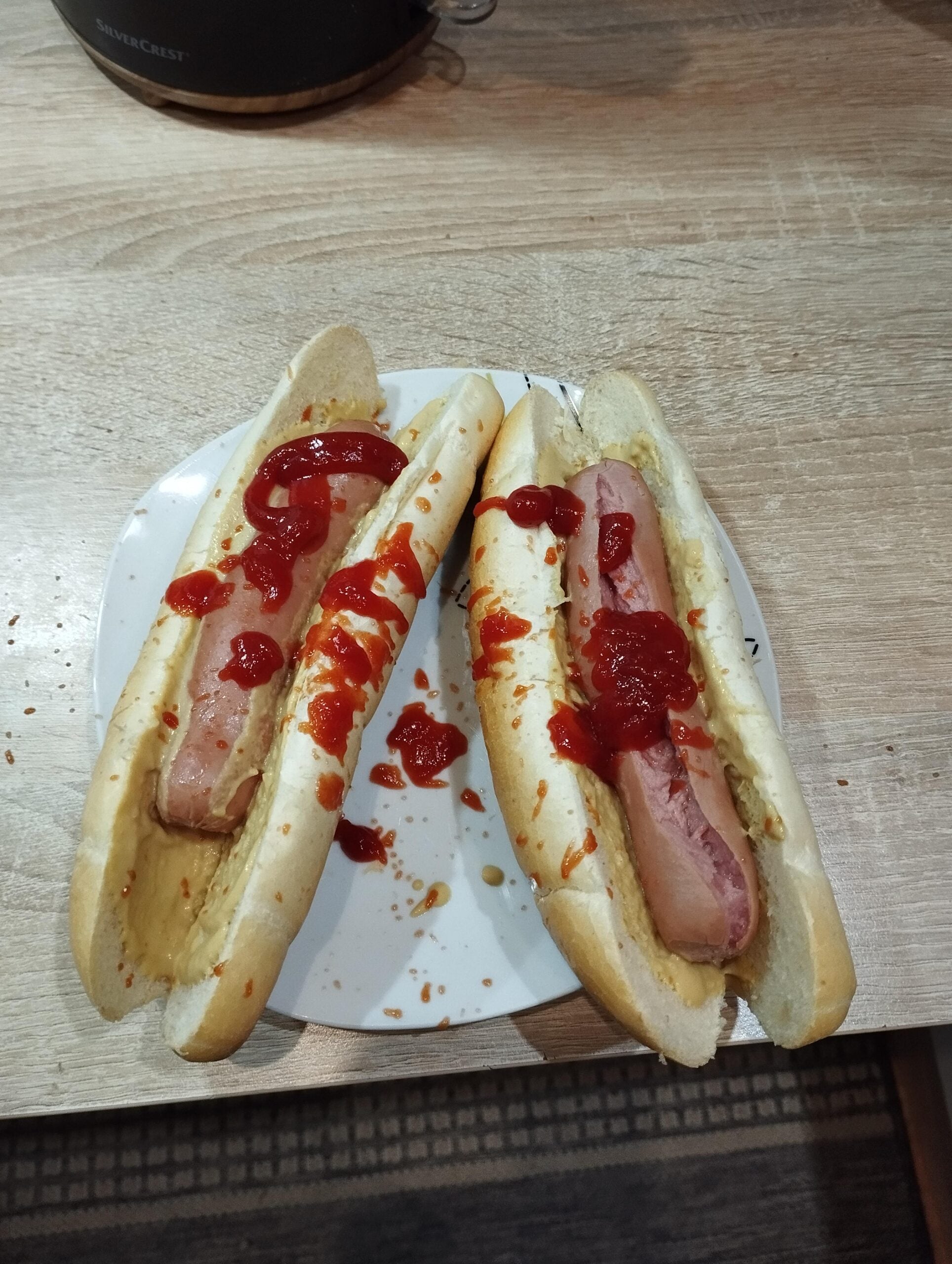 Two hot dogs with ketchup and mustard on a white plate, sitting on a wooden table—evidence of hilarious cooking disasters linger as stray ketchup and crumbs are scattered across the plate and table.