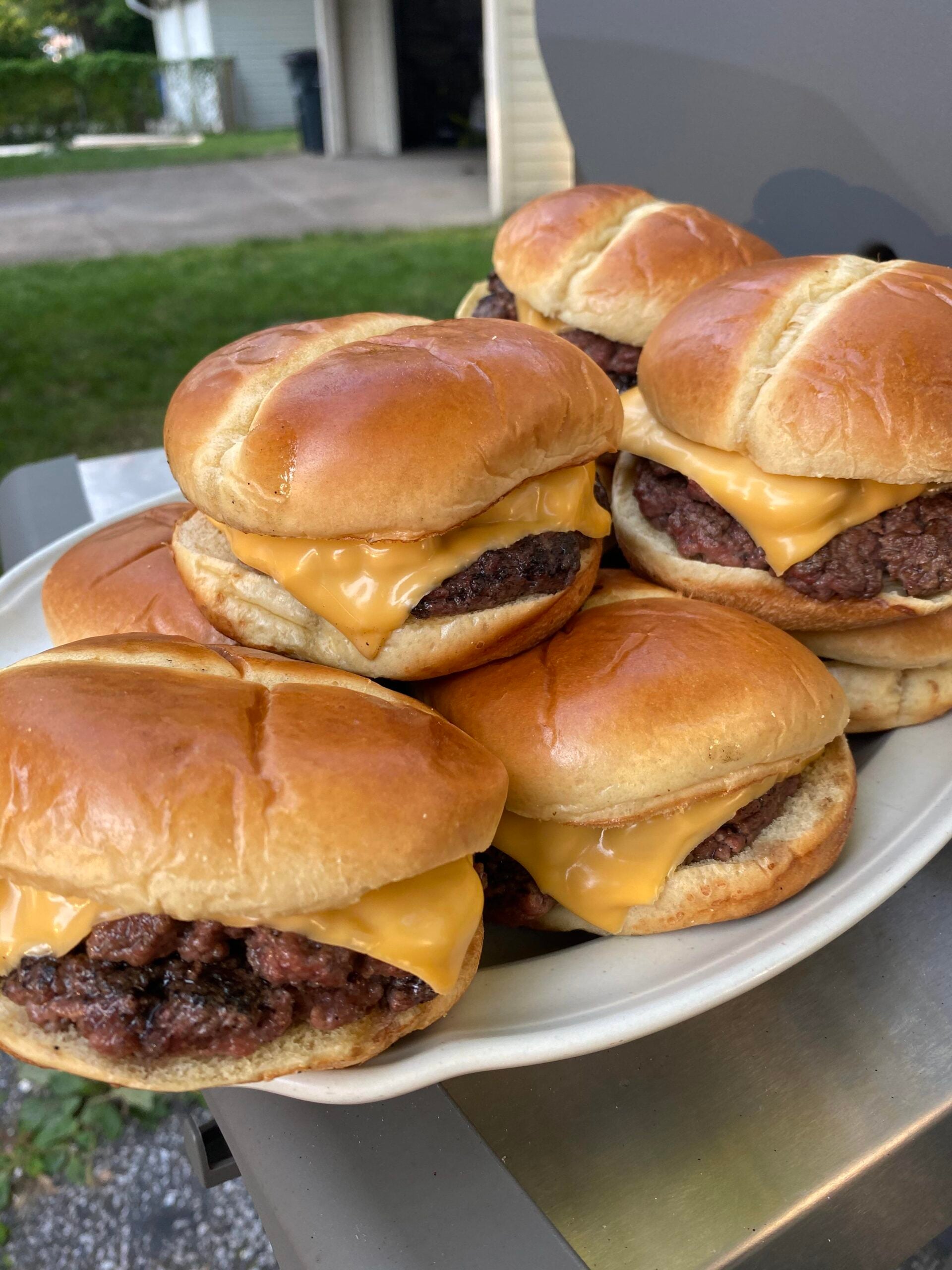 A plate piled with six cheeseburgers, each featuring a slice of melted cheddar cheese on a grilled beef patty and a golden toasted bun, sits outdoors near a grill.