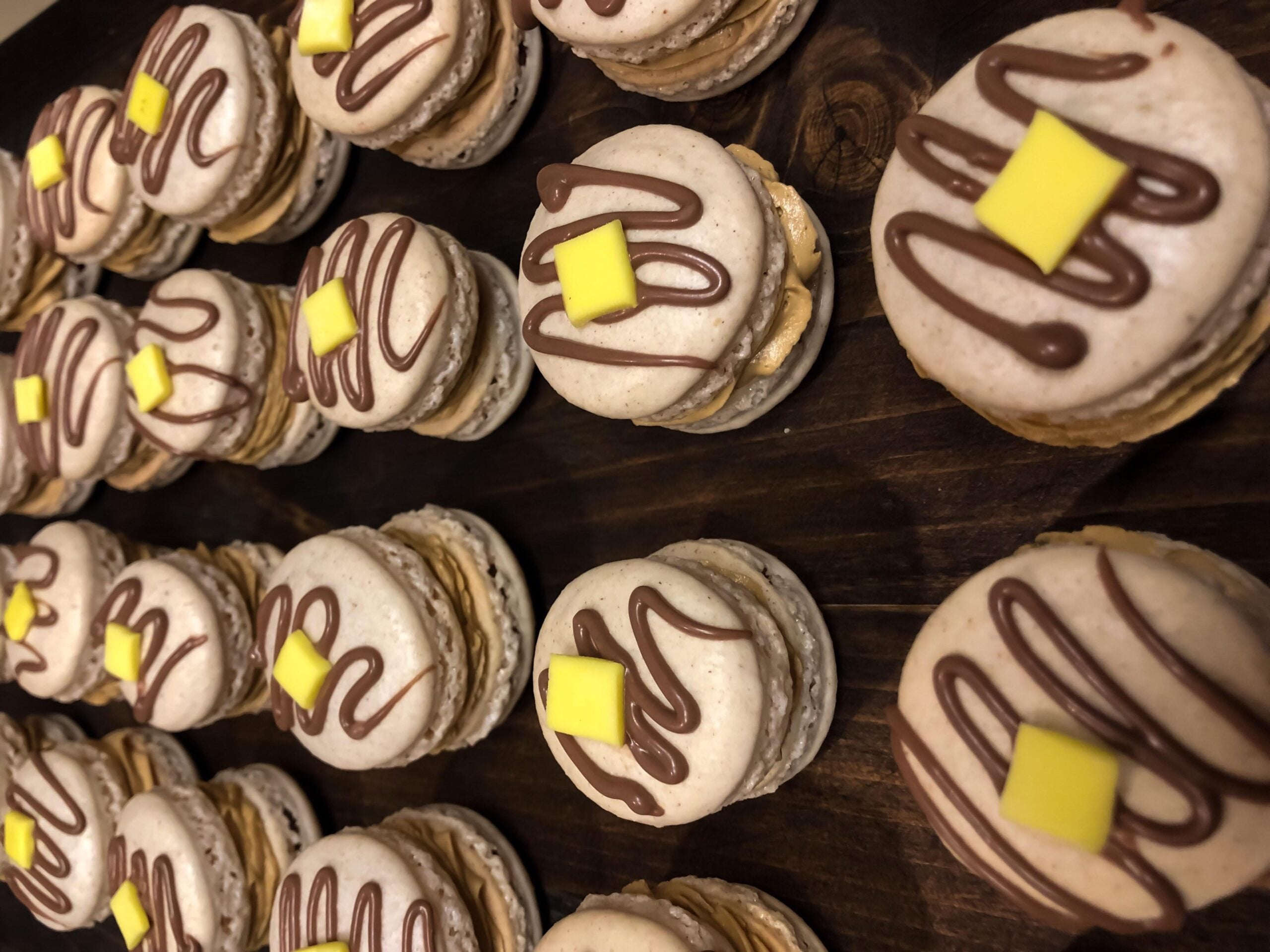 Rows of macarons with chocolate drizzle and a small yellow square on top are arranged on a dark wooden surface, reminiscent of the care and detail found in homemade pancakes.