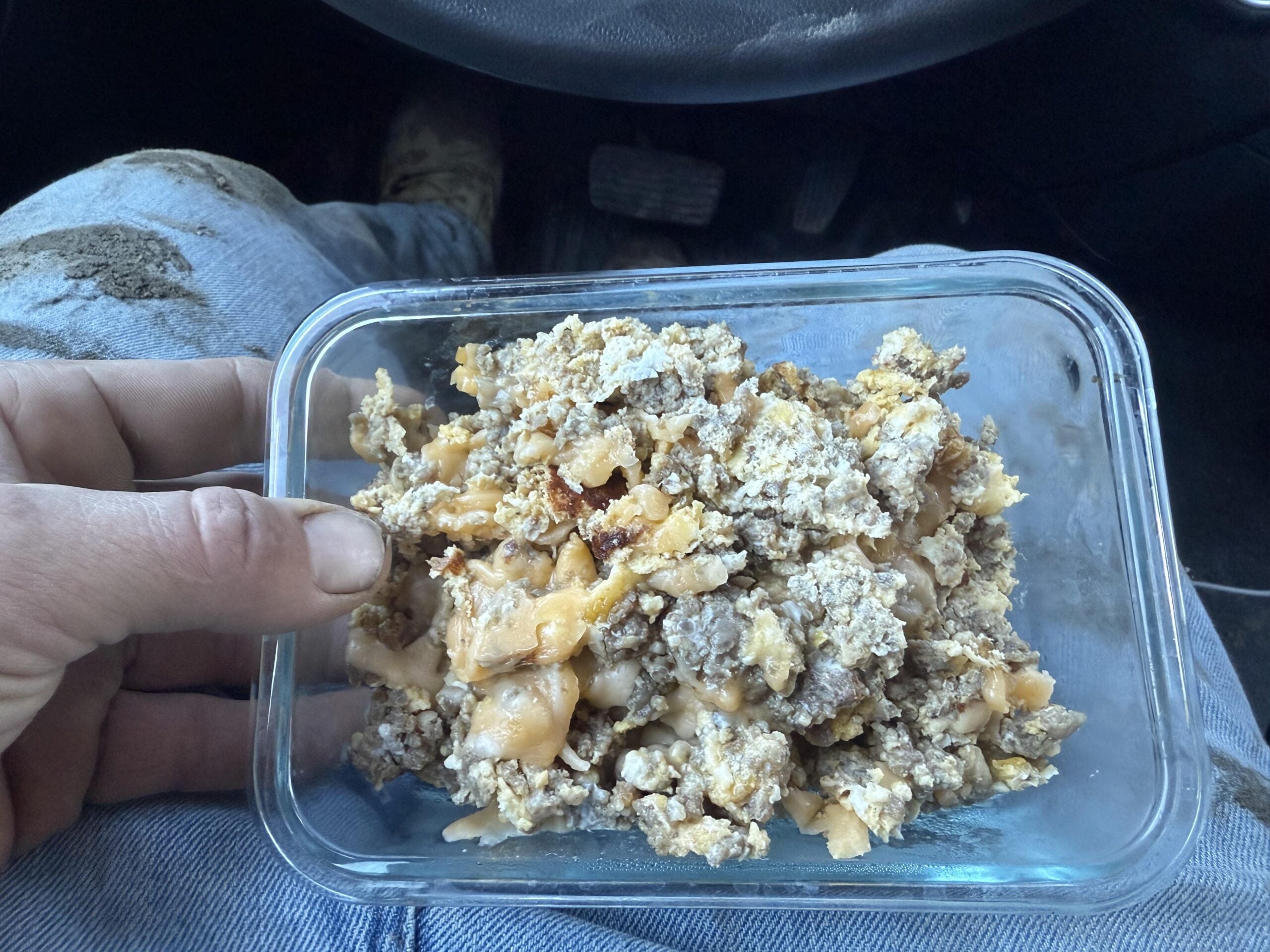 A hand holds a glass container filled with scrambled eggs and ground meat. The person is wearing jeans with dirt stains, and car pedals are visible in the background.