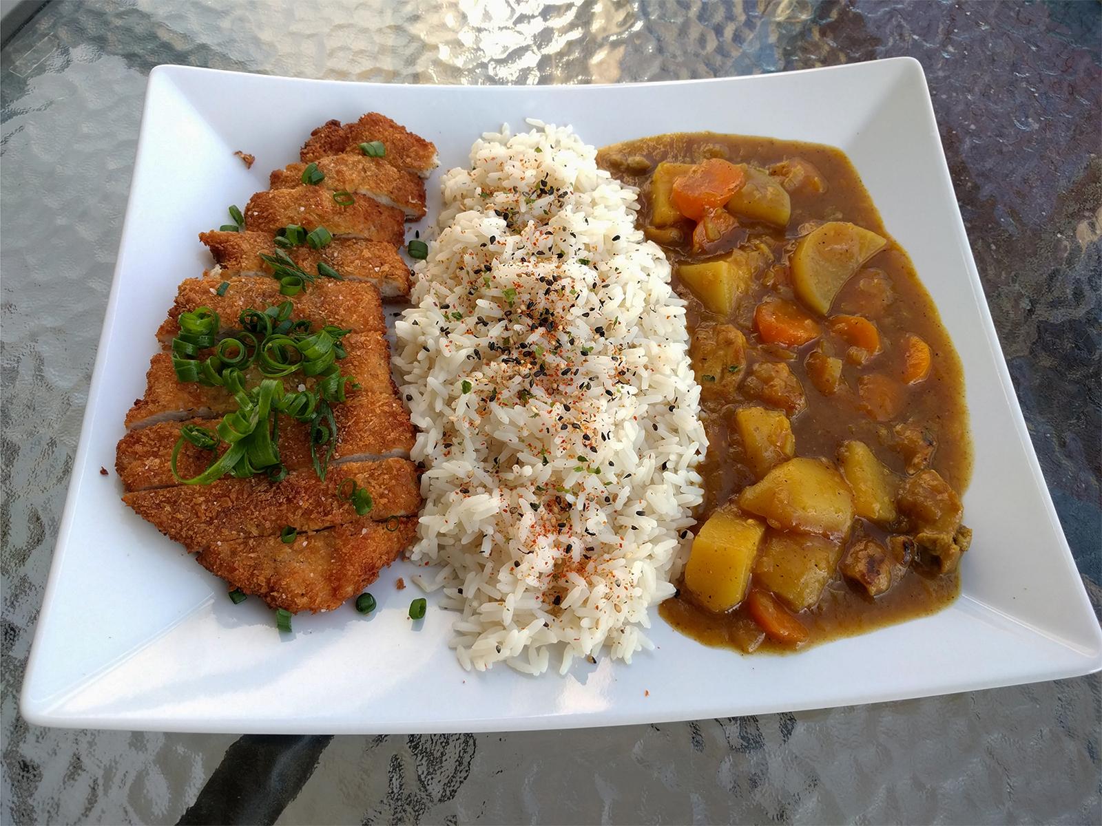 A white plate with sliced breaded cutlet, steamed white rice topped with seasoning, and Japanese curry with potatoes and carrots.