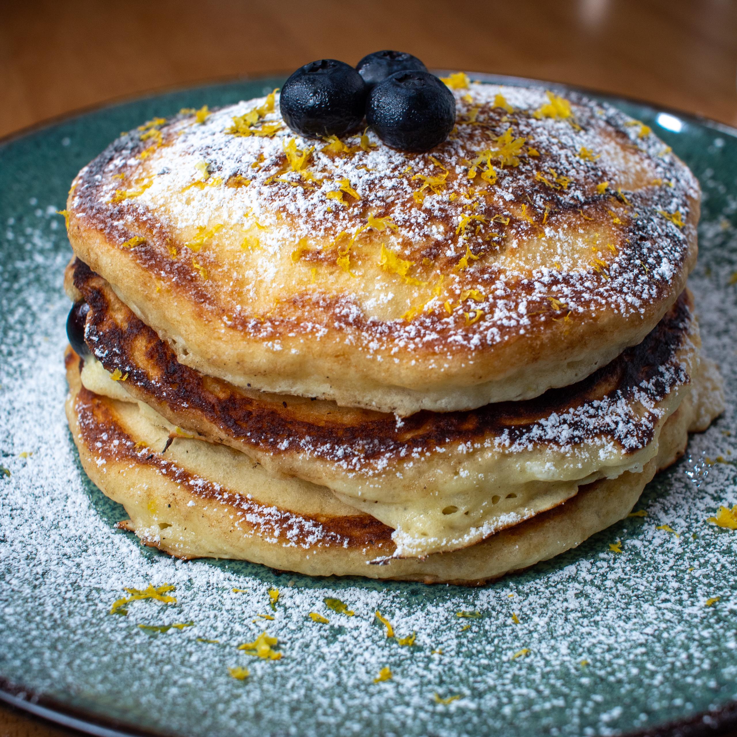 A stack of three fluffy homemade pancakes topped with blueberries, powdered sugar, and orange zest on a green plate.