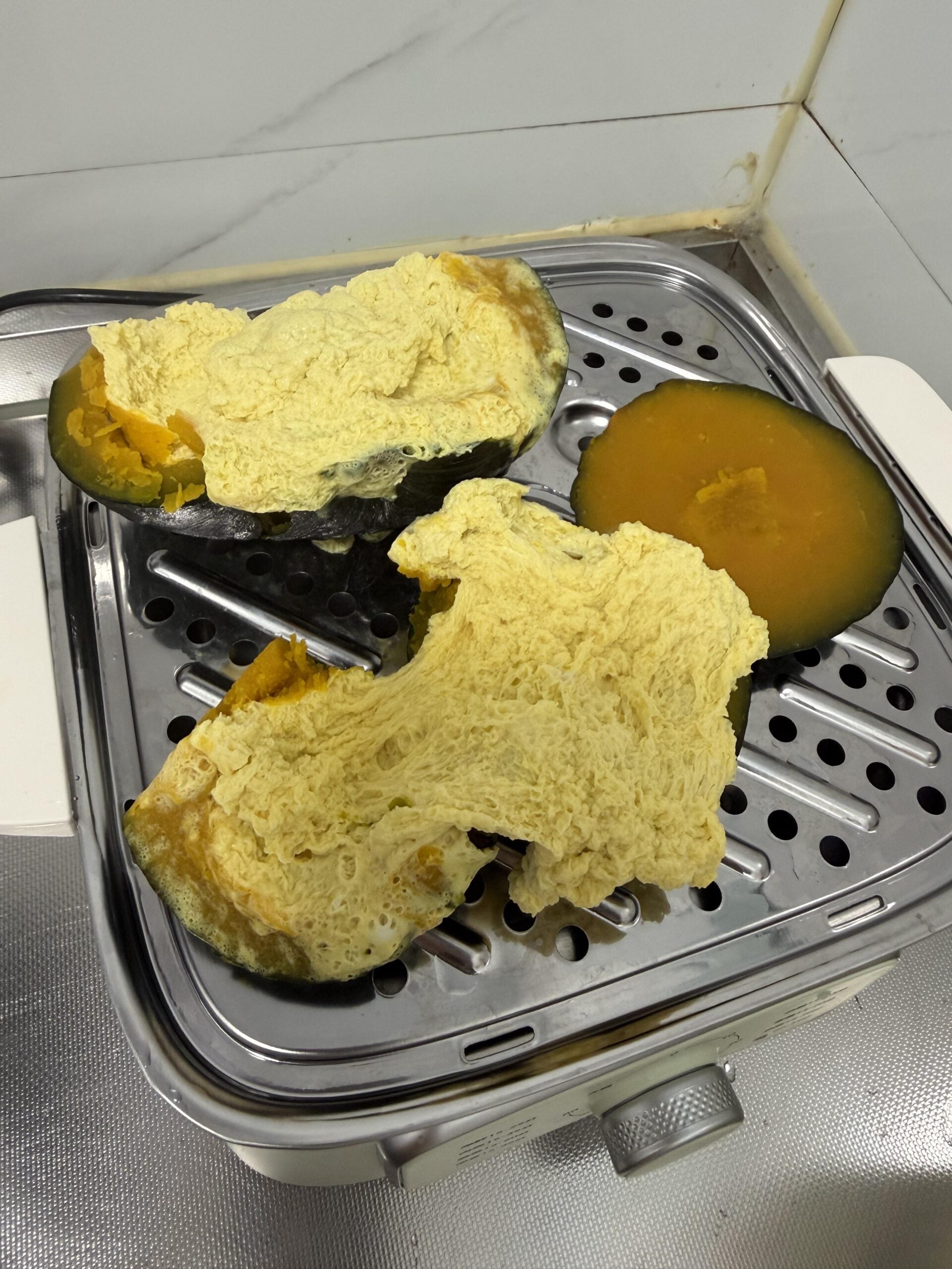 A green-skinned squash is cut open on a metal steamer tray. The inside appears fluffy and spongy, with one slice showing dense orange flesh. The background shows tiled walls and a countertop.