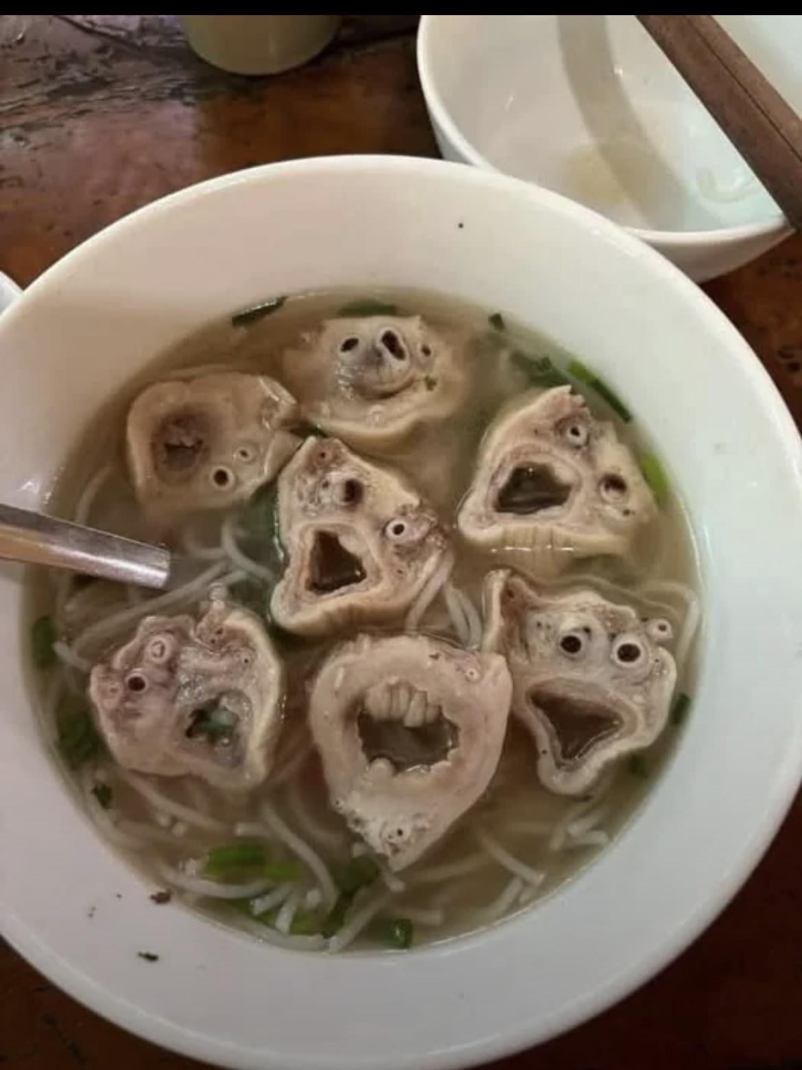 A bowl of noodle soup contains several meat pieces with holes that resemble surprised or shocked cartoon faces, along with chopped green onions.