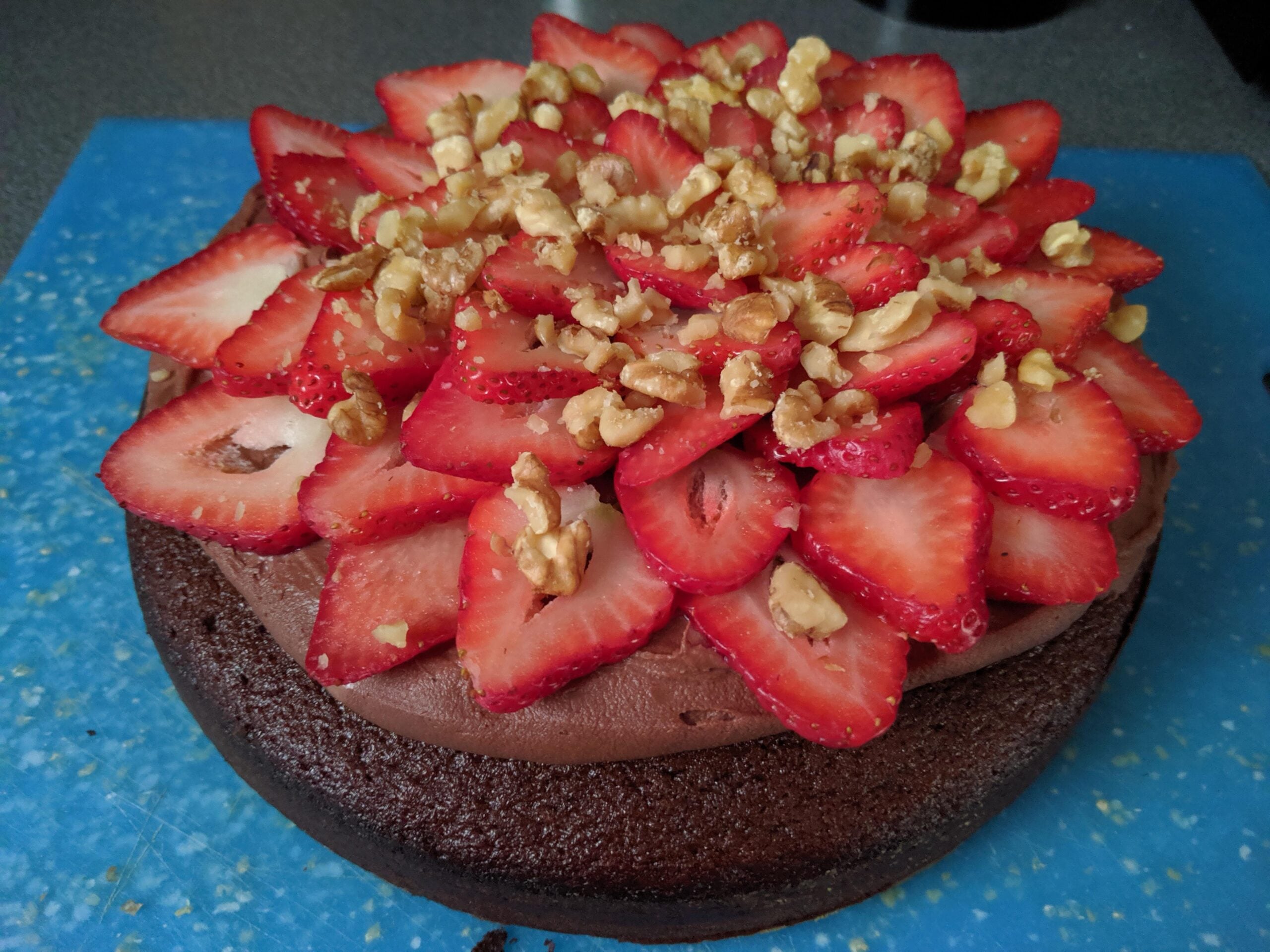 A delectable chocolate cake topped with rich chocolate frosting, sliced fresh strawberries arranged in layers, and chopped walnuts, all on a blue cutting board—a perfect example of homemade desserts.