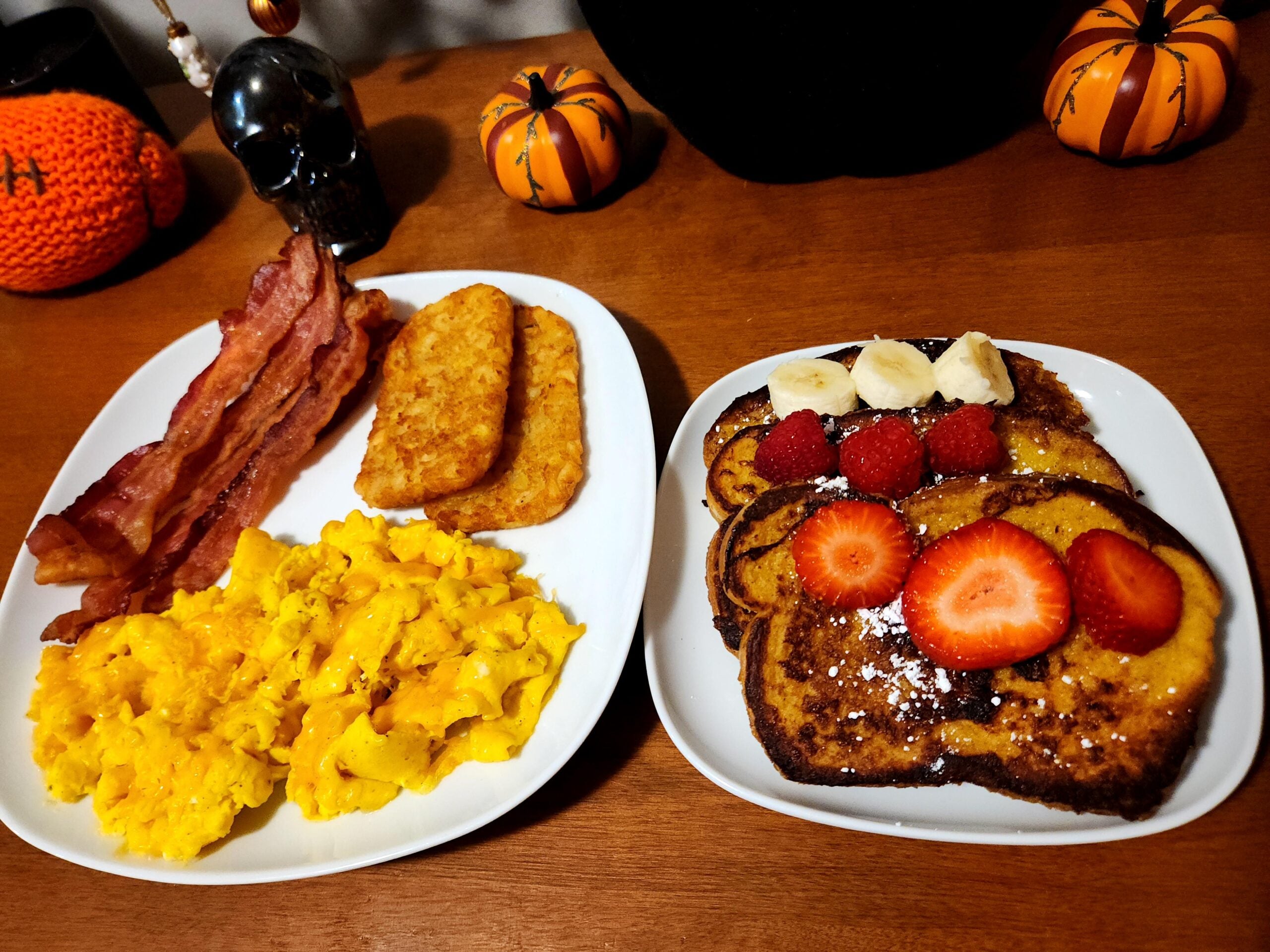 Two plates: One with scrambled eggs, bacon, and hash browns. The other with French toast topped with strawberries, raspberries, banana slices, and powdered sugar. Halloween pumpkin decor in the background.
