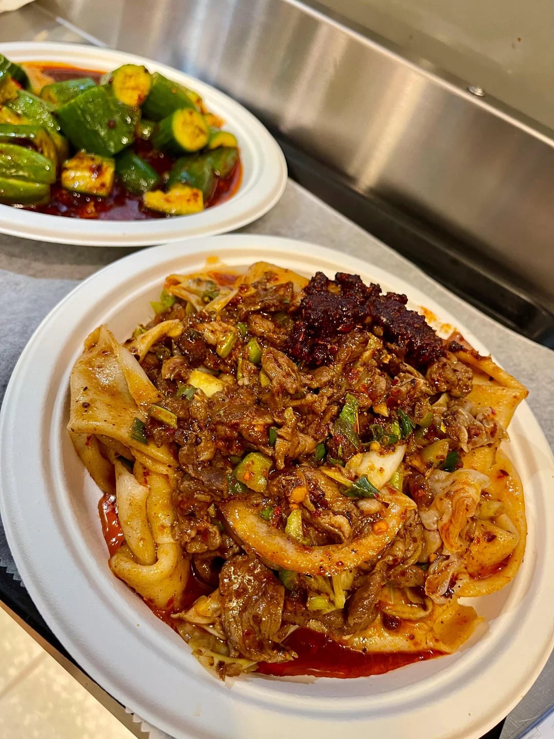 A plate of spicy hand-pulled noodles with beef, vegetables, and chili paste, accompanied by a side dish of marinated cucumber slices in a red chili sauce on a nearby plate.