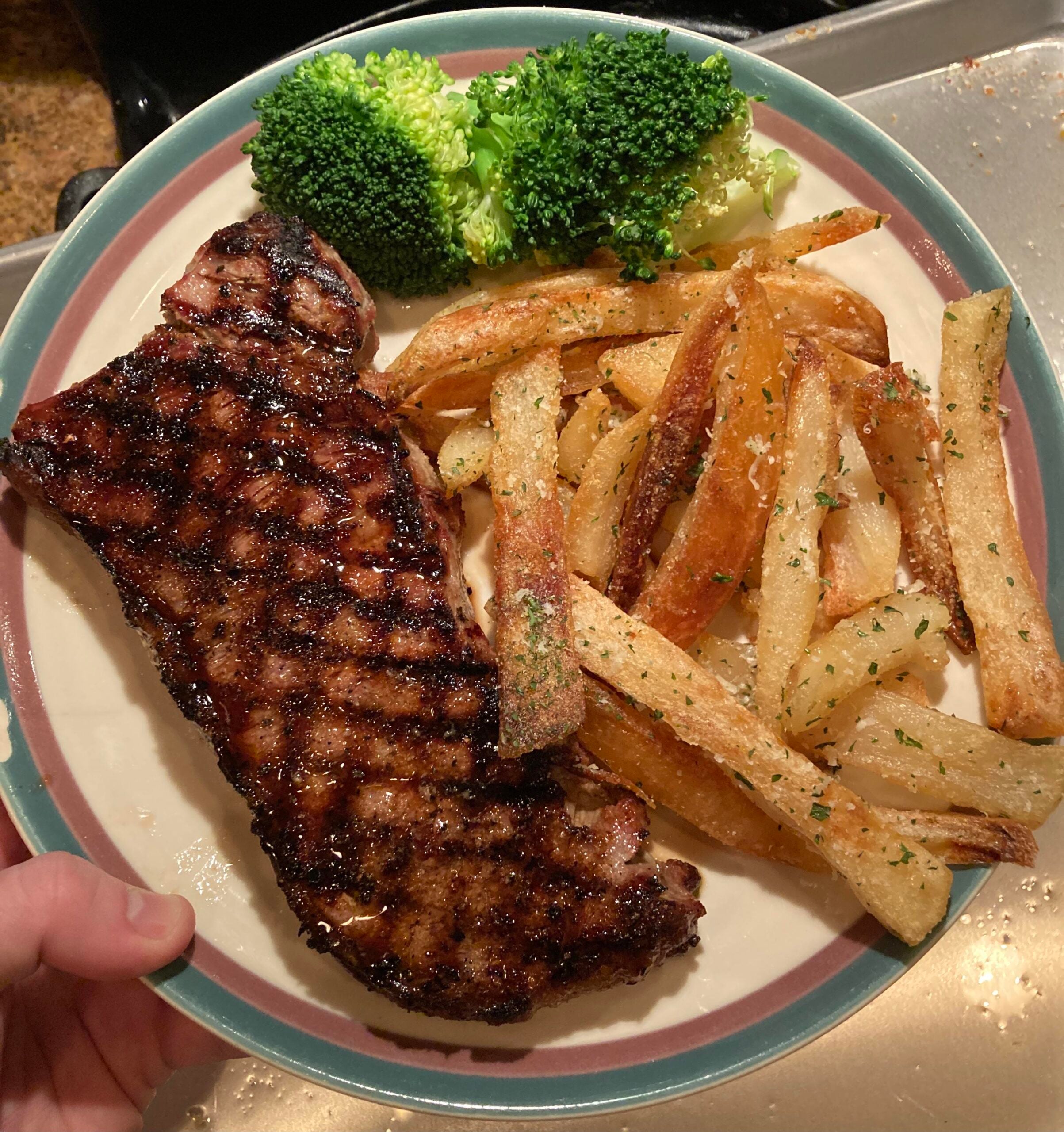 A plate with a grilled steak, seasoned French fries, and steamed broccoli. A hand is holding the plate over a kitchen counter and sink.