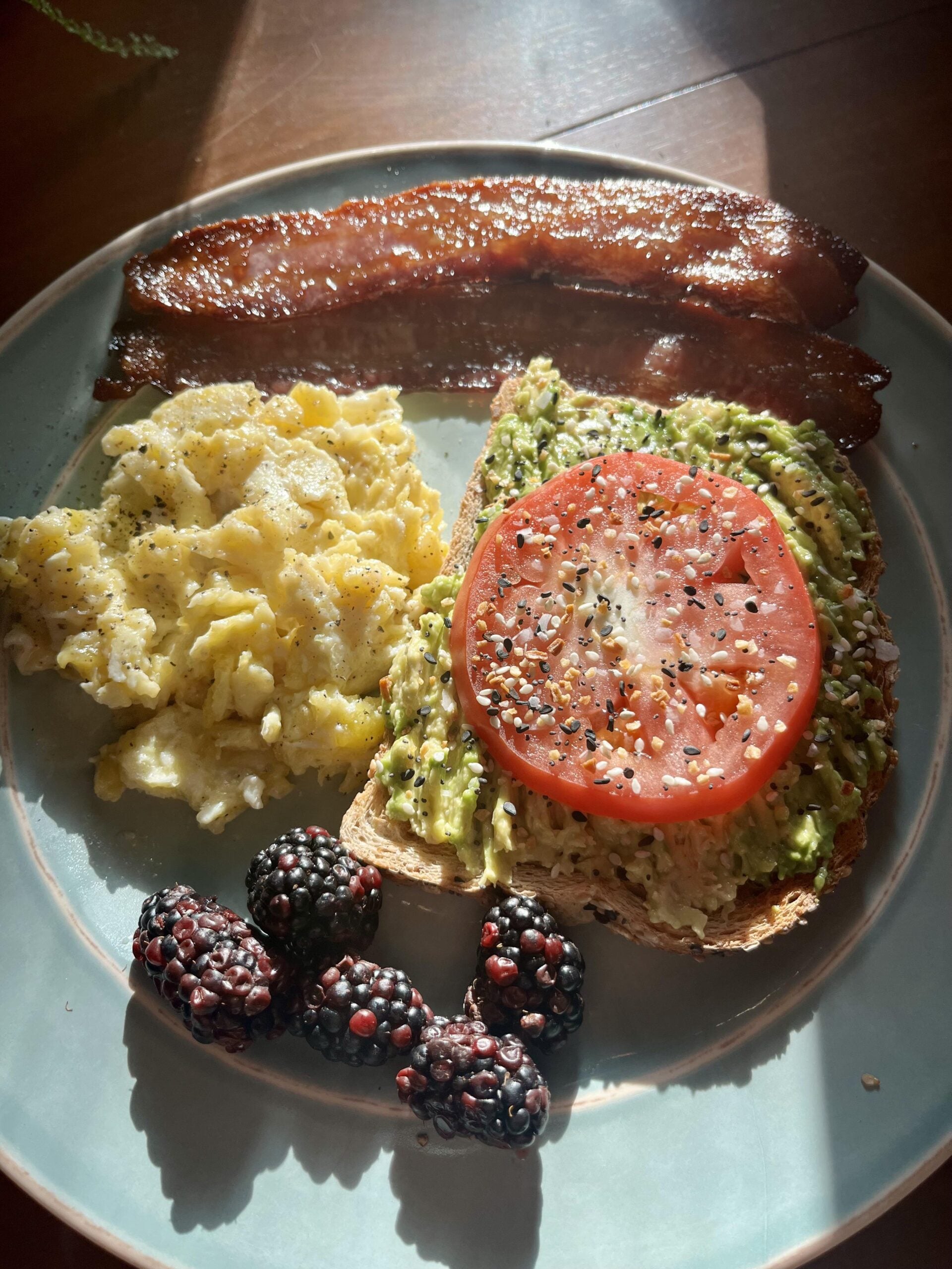 A plate with scrambled eggs, two strips of bacon, avocado toast topped with tomato and seasoning, and four blackberries, all illuminated by sunlight.
