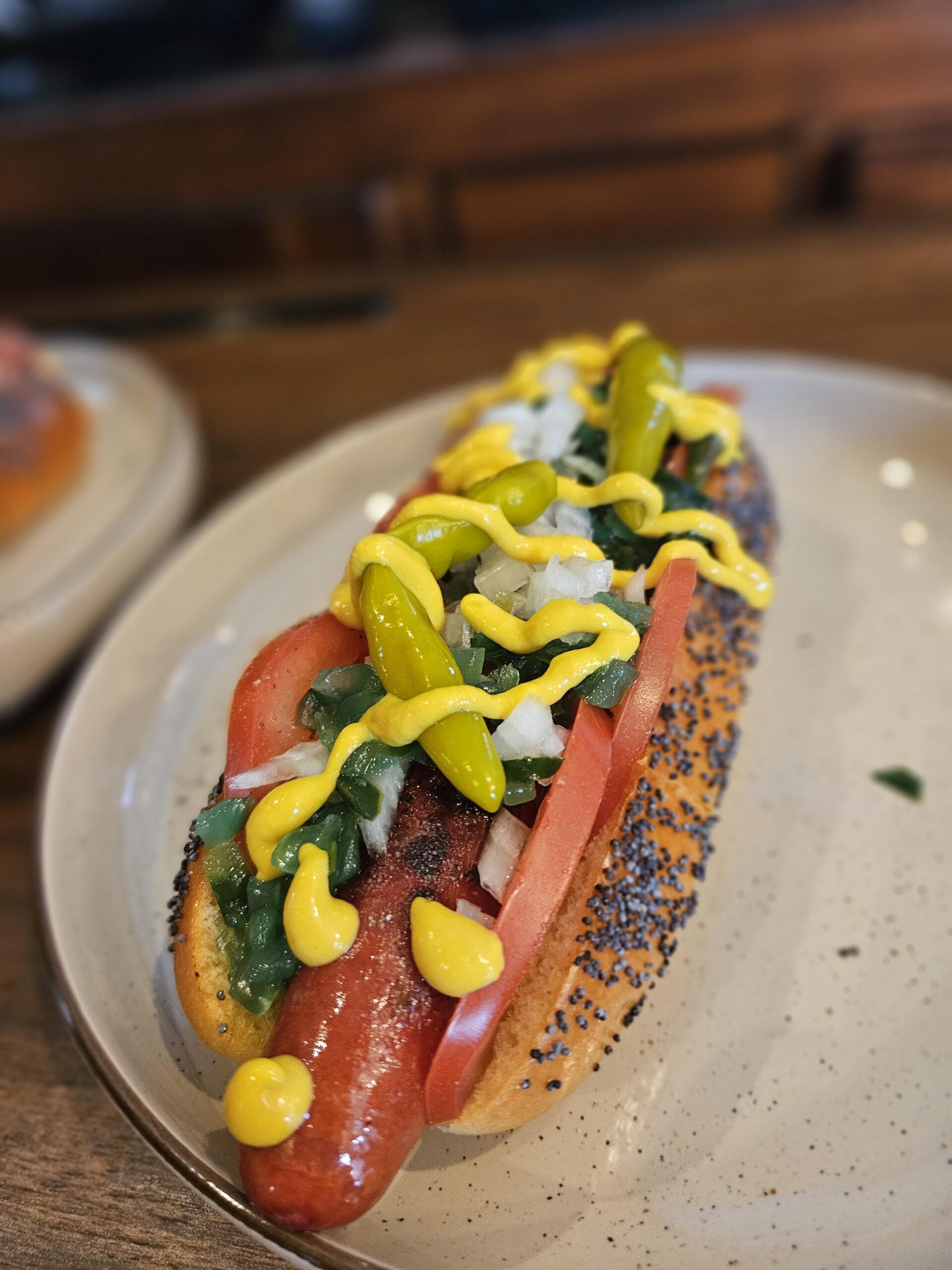 A Chicago-style hot dog in a poppy seed bun topped with mustard, onions, relish, tomato slices, sport peppers, and a pickle spear, served on a white plate.