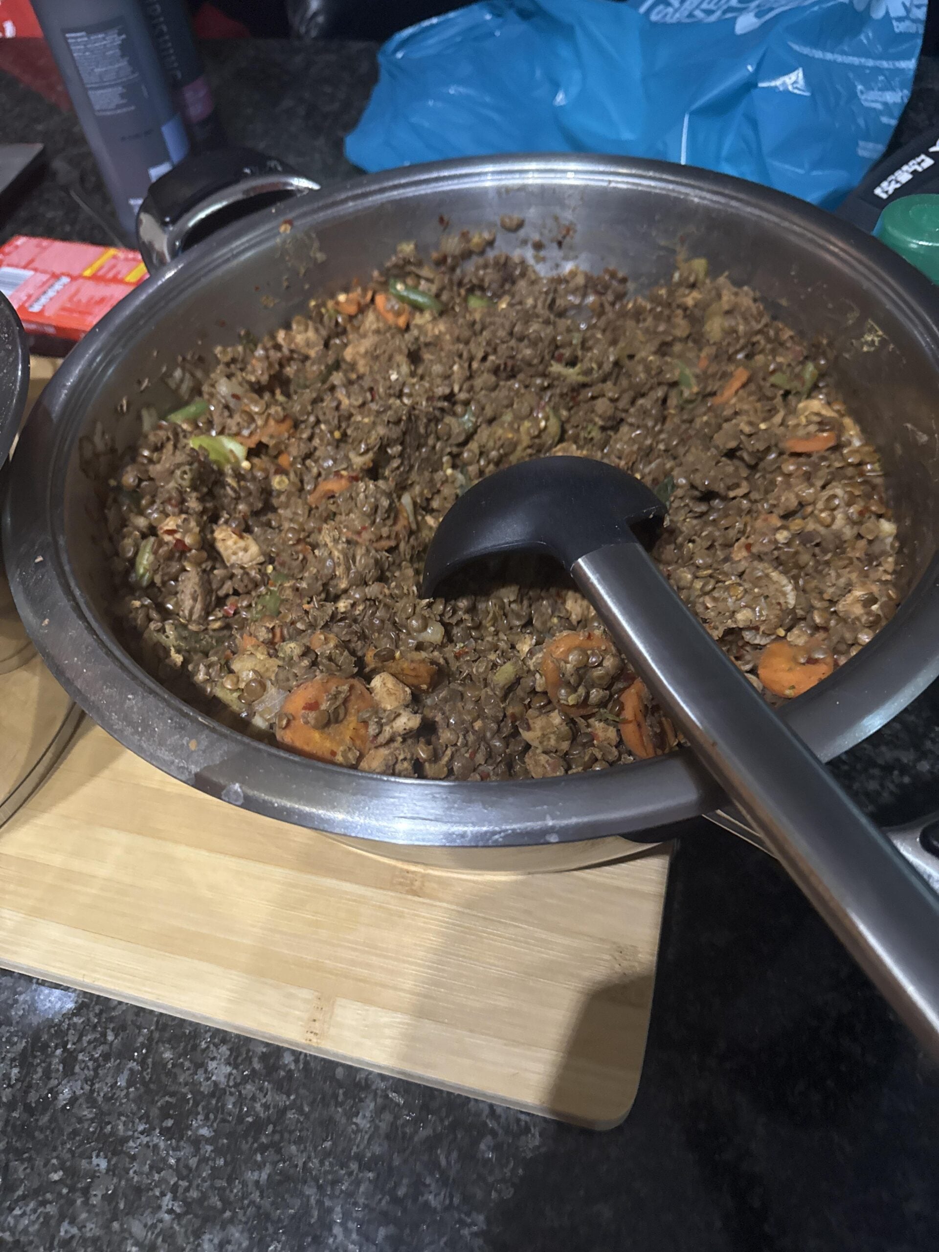 A large pot filled with a cooked mixture of lentils, vegetables, and sauce sits on a wooden board, with a black ladle resting inside.
