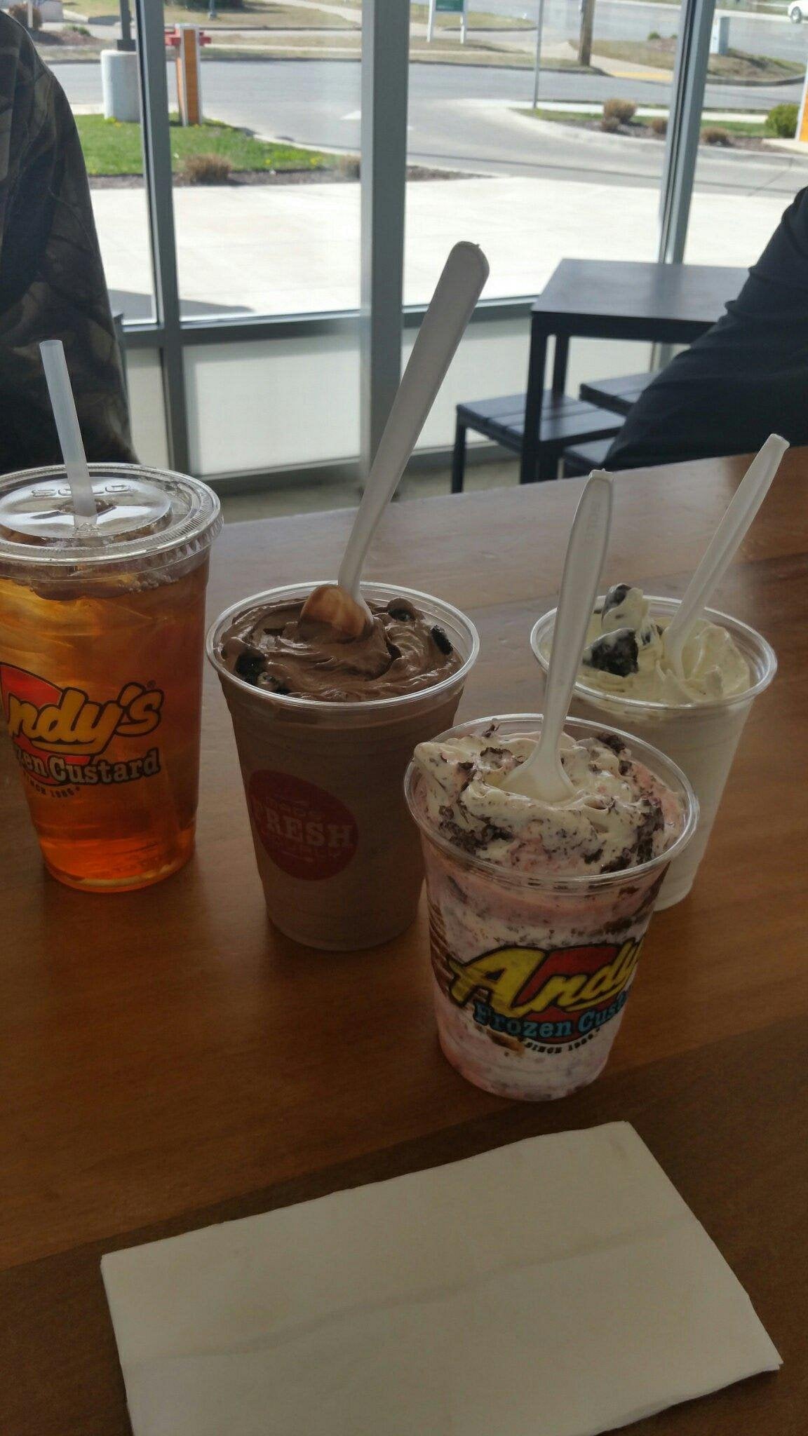 Four frozen custard desserts and an iced drink on a wooden table inside a brightly lit restaurant belonging to one of the beloved regional fast-food chains, with napkins and plastic spoons, and windows showing a street outside.