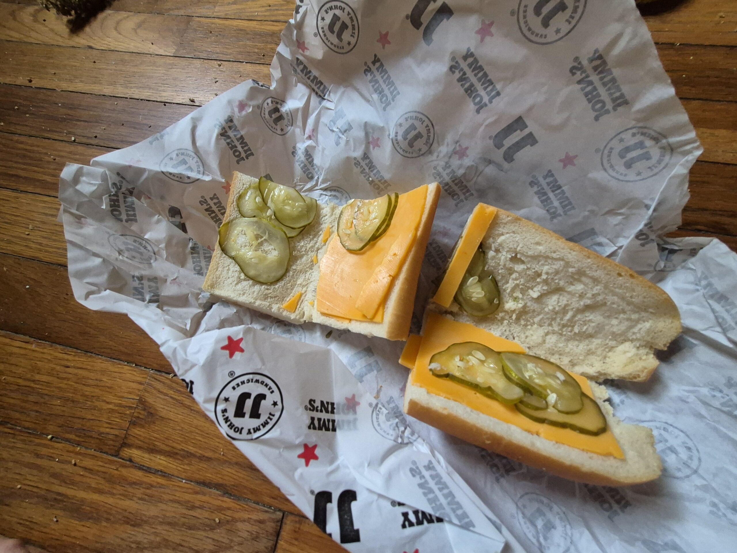 A sandwich from Jimmy John's with cheese and pickles on white bread, cut in two halves, sits on branded sandwich paper on a wooden floor.