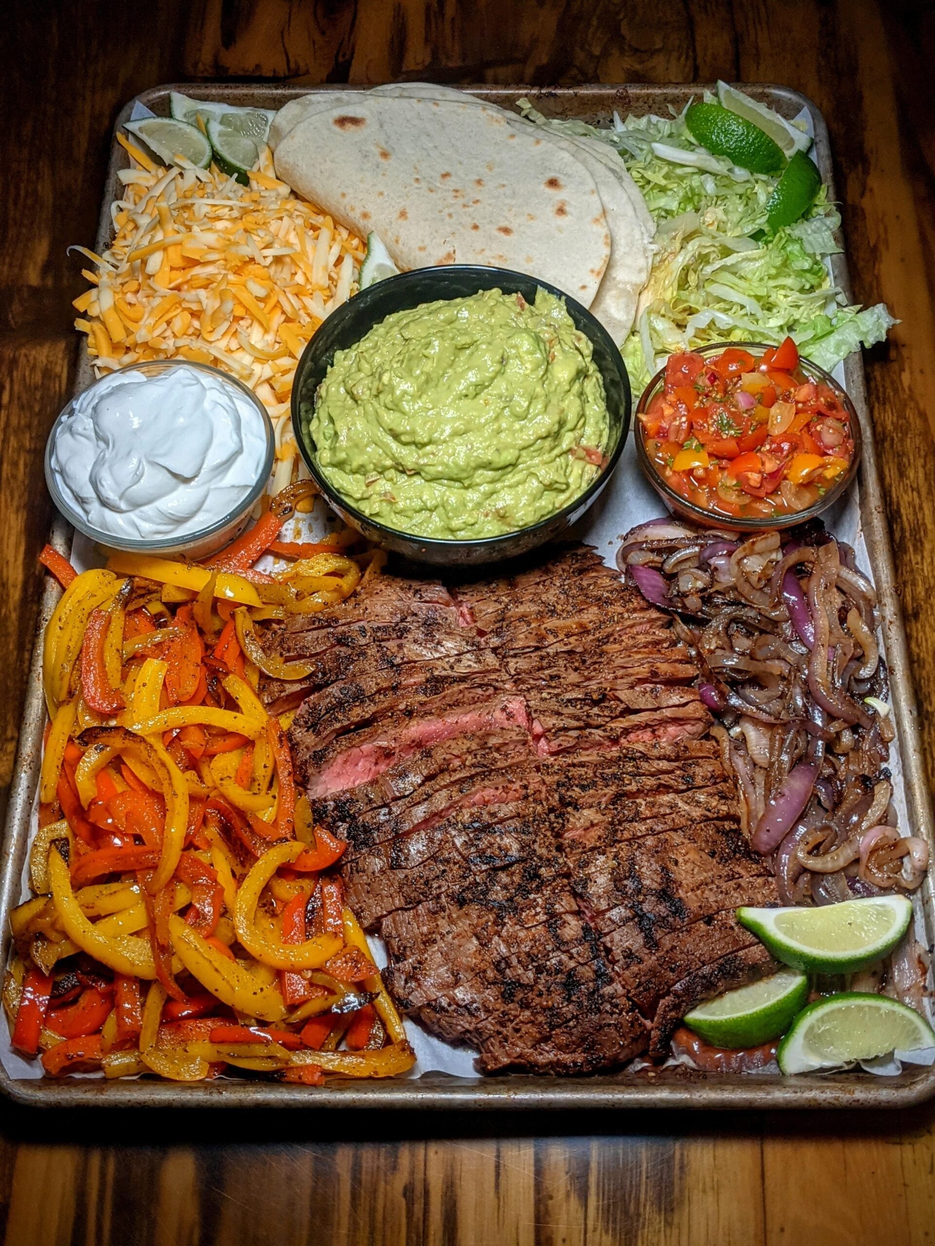 A tray with grilled steak, sautéed peppers and onions, shredded cheese, tortillas, lettuce, guacamole, sour cream, salsa, and lime wedges, arranged for fajitas.