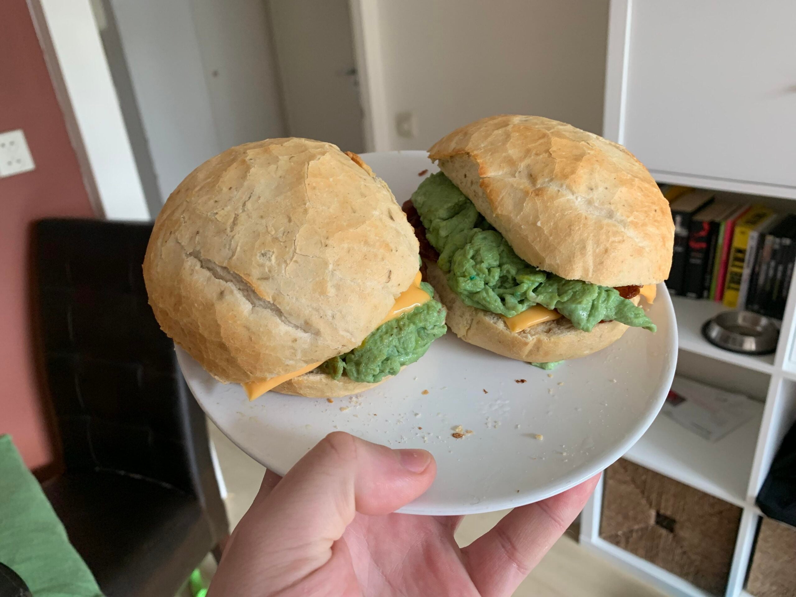 A hand holds a white plate with two sandwiches. Each sandwich has a round bun, a slice of yellow cheese, and a green filling. A bookshelf and a chair are visible in the background.