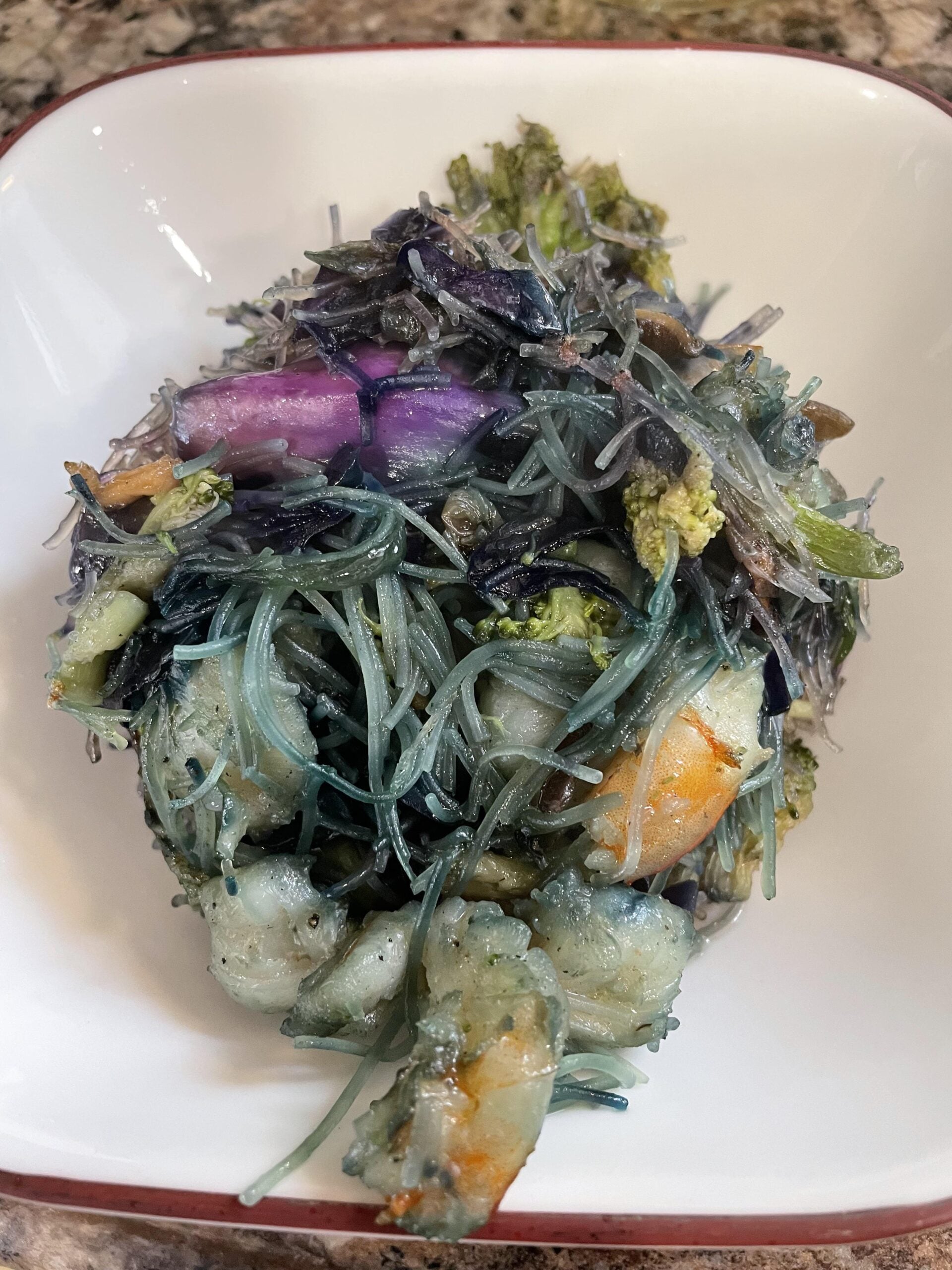 A white bowl filled with glass noodles, shrimp, broccoli, and purple vegetables, all mixed together in a colorful stir-fry.