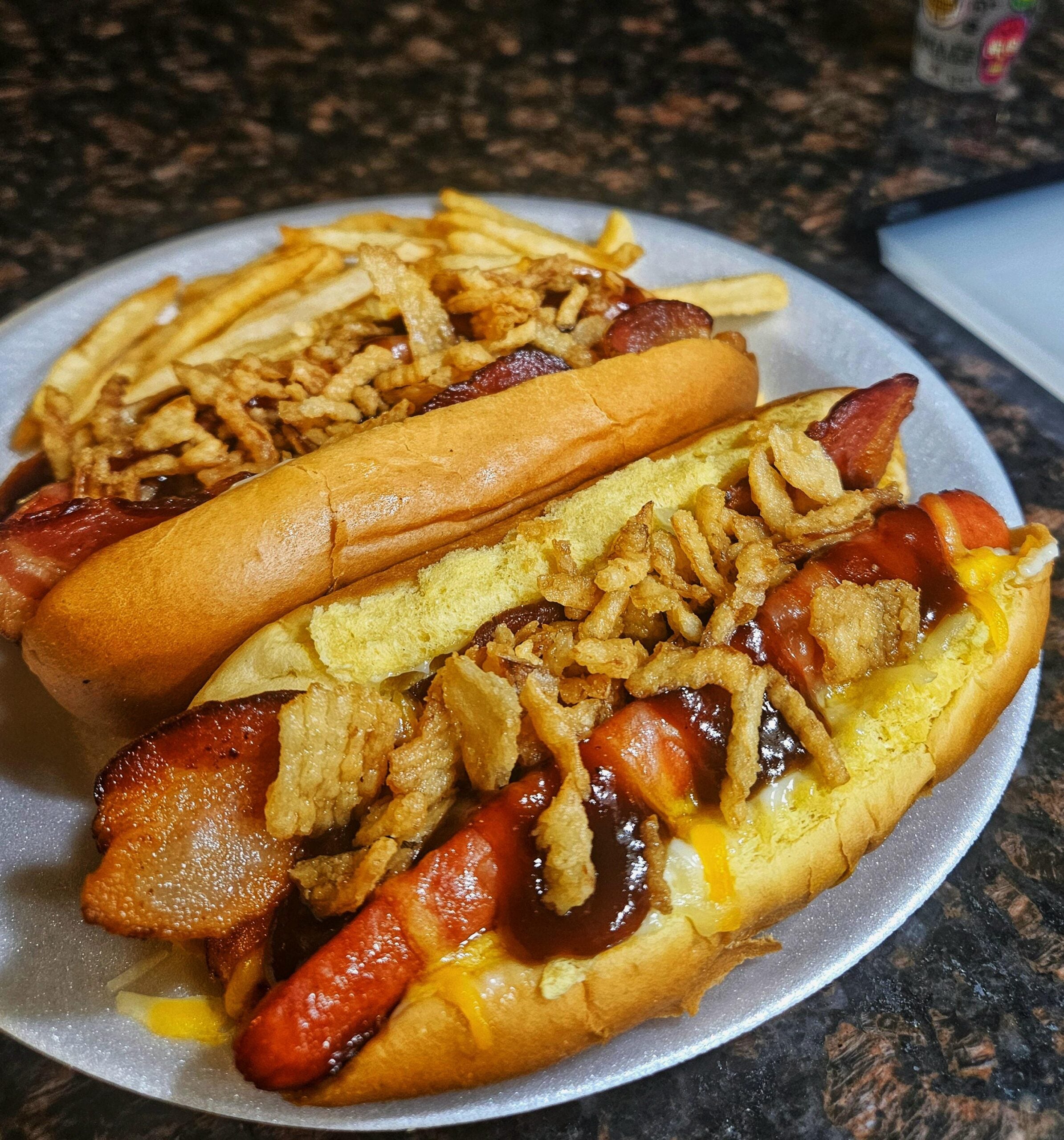 Two hot dogs topped with crispy onions, bacon, mustard, and barbecue sauce sit on a plate next to a pile of thin French fries on a dark granite countertop.