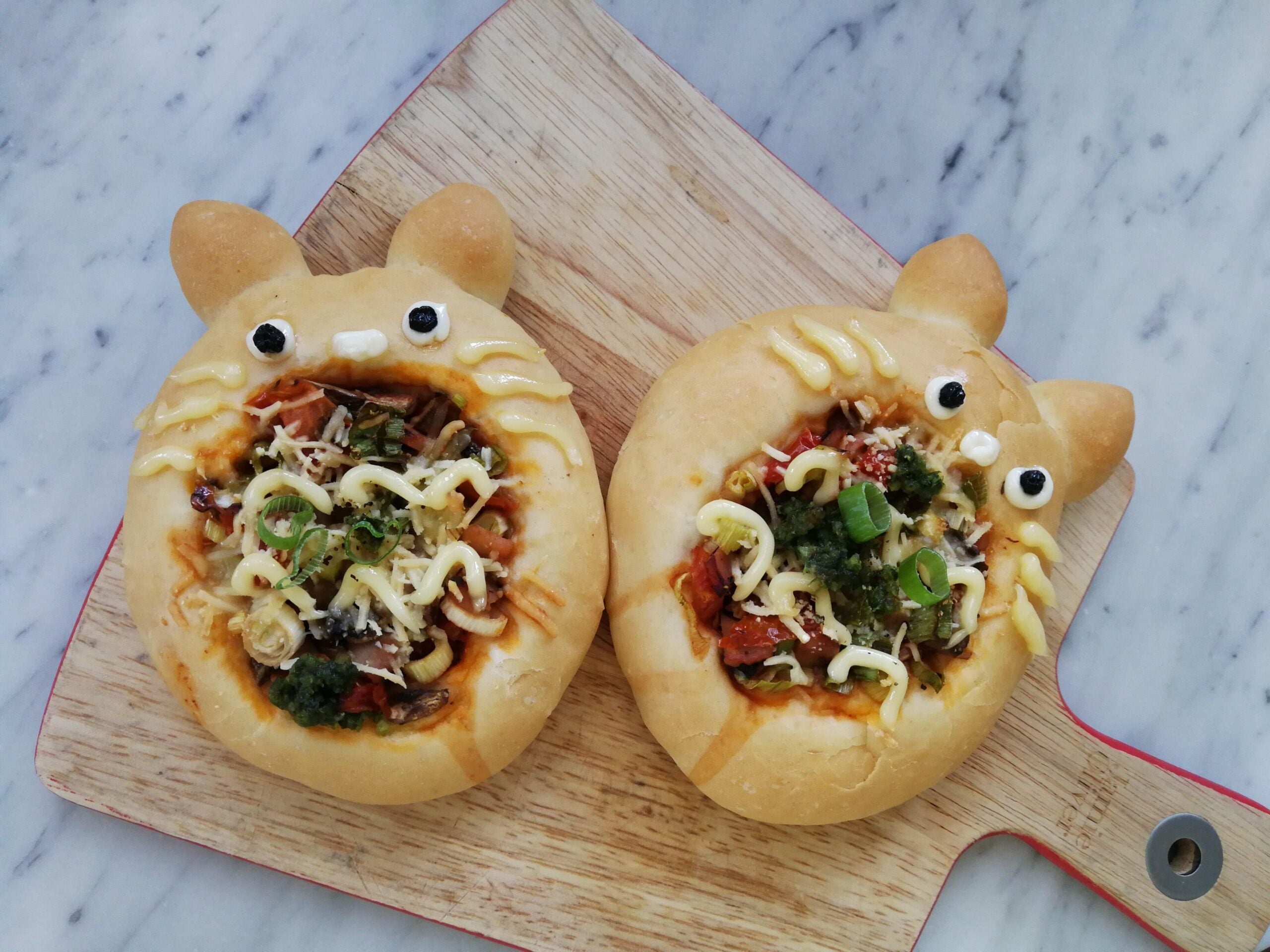 Two round breads decorated to look like cartoon cats, filled with savory toppings like vegetables, cheese, and sauce, placed on a wooden cutting board on a marble surface.