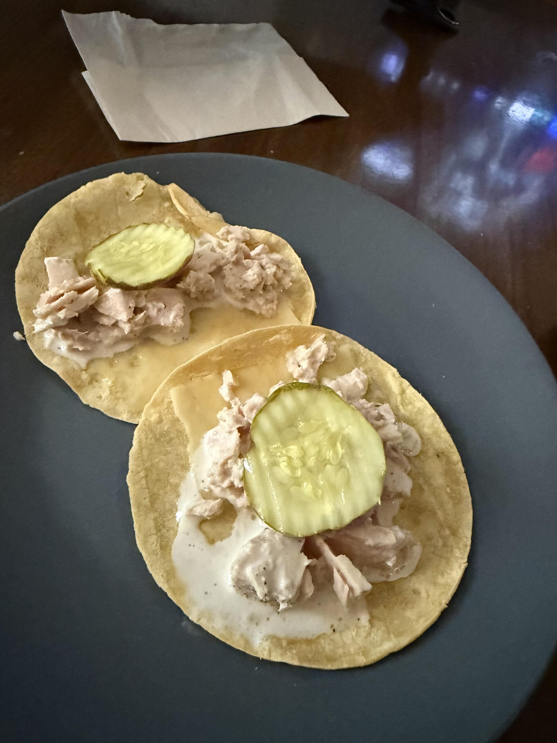 Two corn tortillas topped with pieces of chicken, creamy white sauce, and a pickle slice on each, served on a gray plate with a napkin in the background.