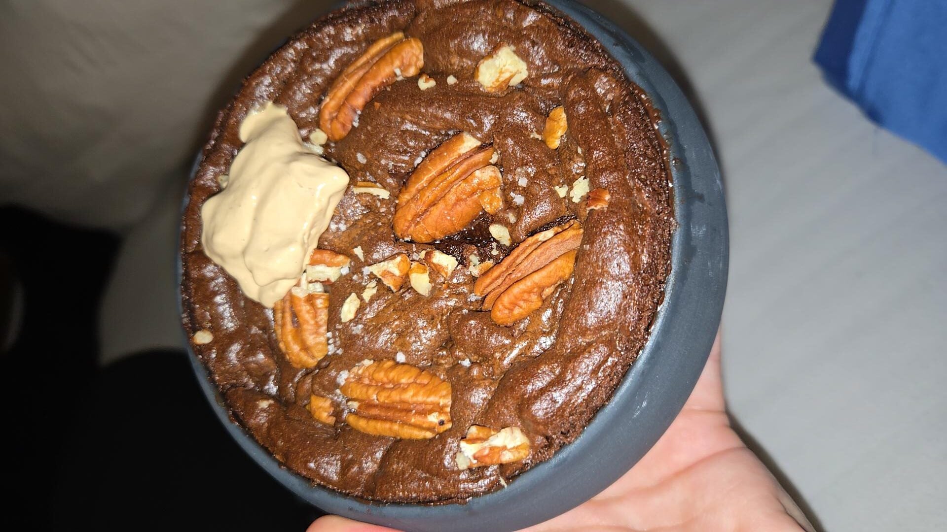 A hand holds a small bowl with a baked chocolate dessert topped with pecan halves and a dollop of creamy spread. The background is slightly blurred.