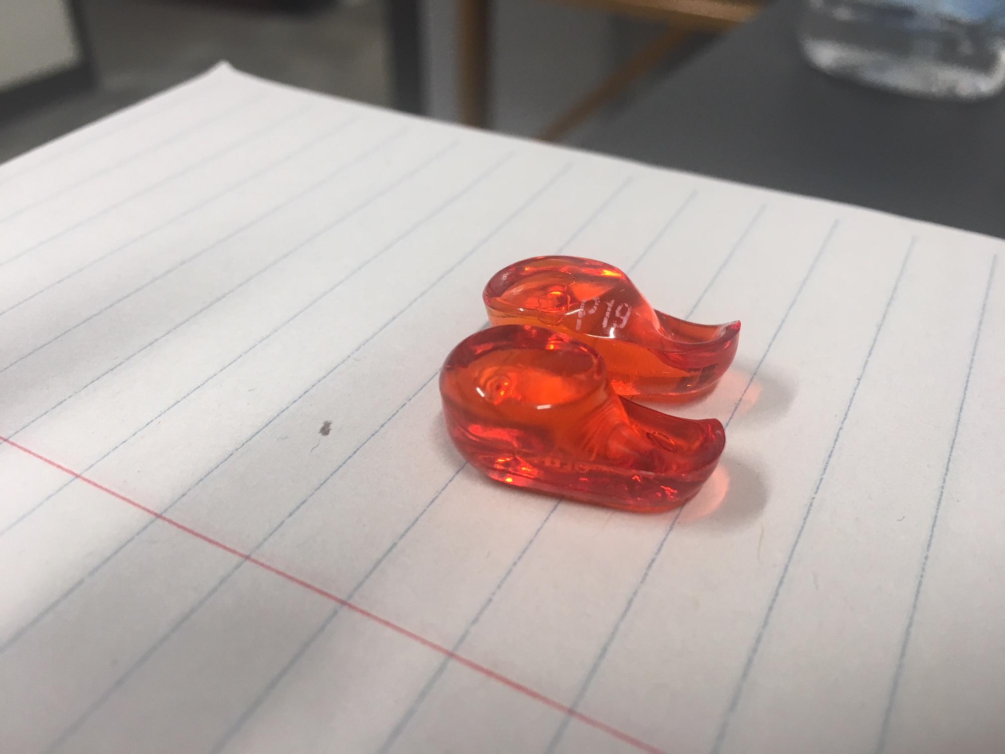 Two small, translucent red plastic objects shaped like shoes sit on a lined notebook page, with a desk and a water bottle blurred in the background.