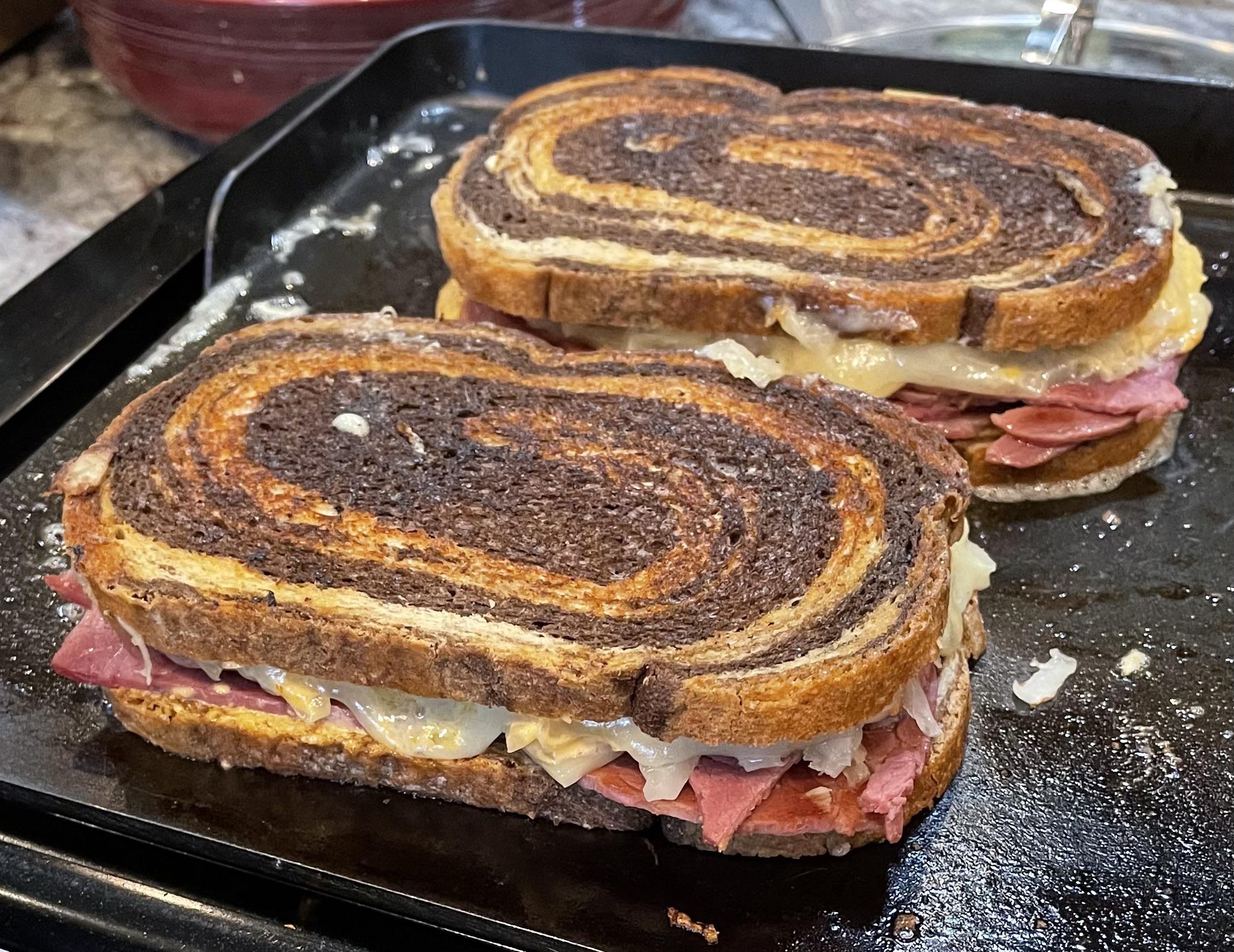 Two Reuben sandwiches with swirled rye bread, corned beef, melted cheese, and sauerkraut are being grilled on a stovetop griddle.