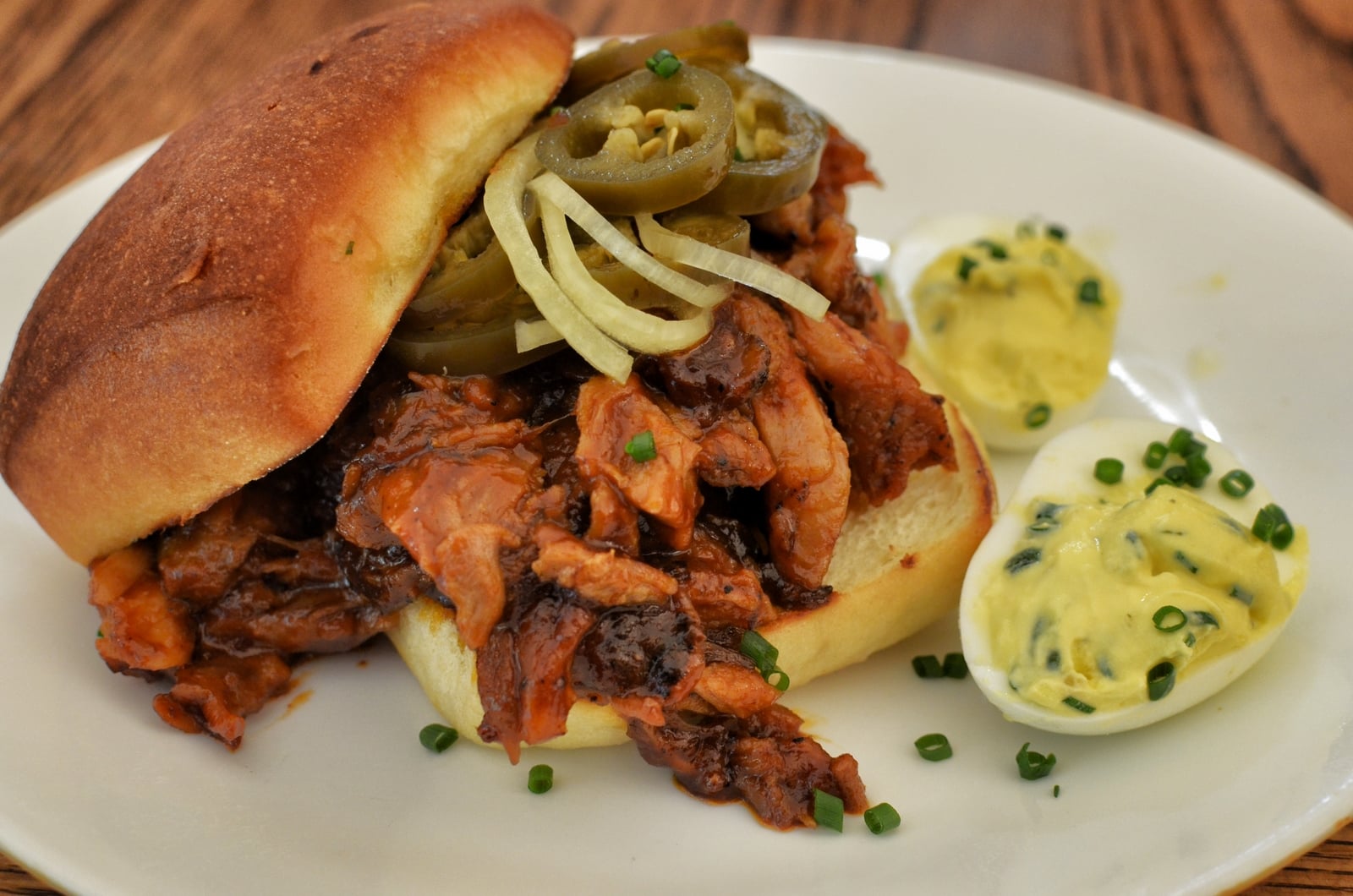 A pulled pork sandwich topped with jalapeños and onions sits on a plate next to two deviled eggs garnished with chopped chives.