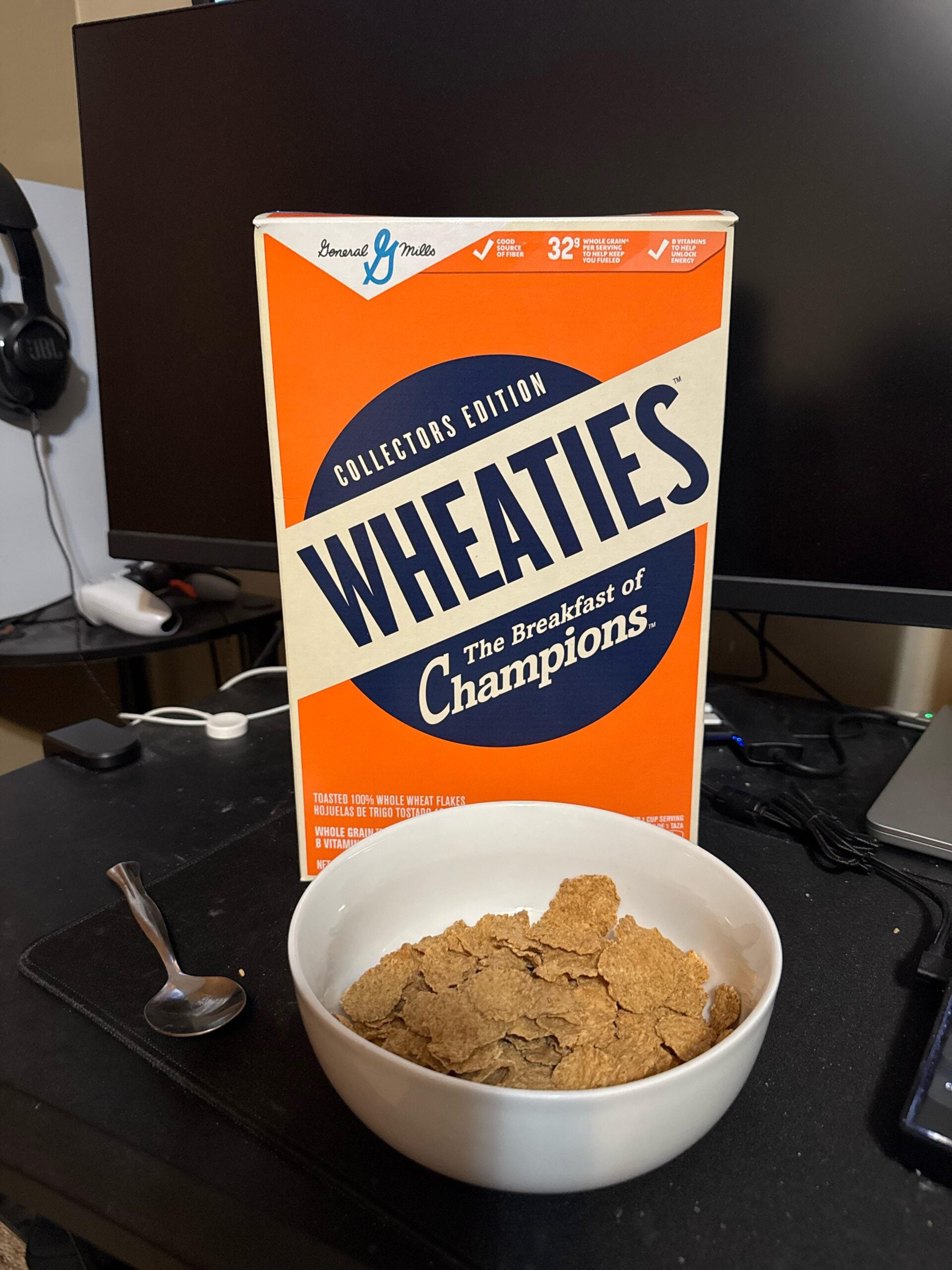 A bowl of Wheaties cereal sits in front of a Wheaties cereal box on a desk, with a spoon to the side. A computer monitor, headphone, and other electronics are visible in the background.