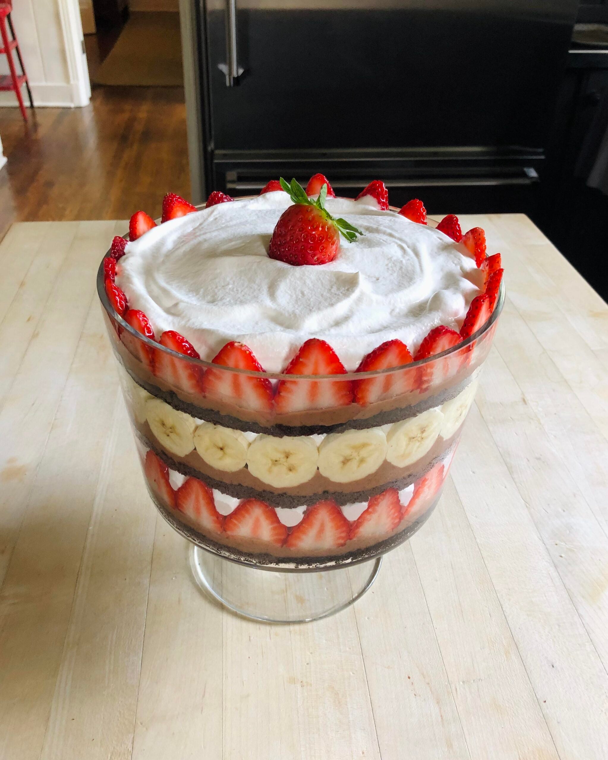 A delightful layered trifle in a glass bowl featuring strawberries, banana slices, chocolate cake, and whipped cream—one of the most irresistible homemade Christmas treats—topped with a whole strawberry and garnished with sliced strawberries around the edge.