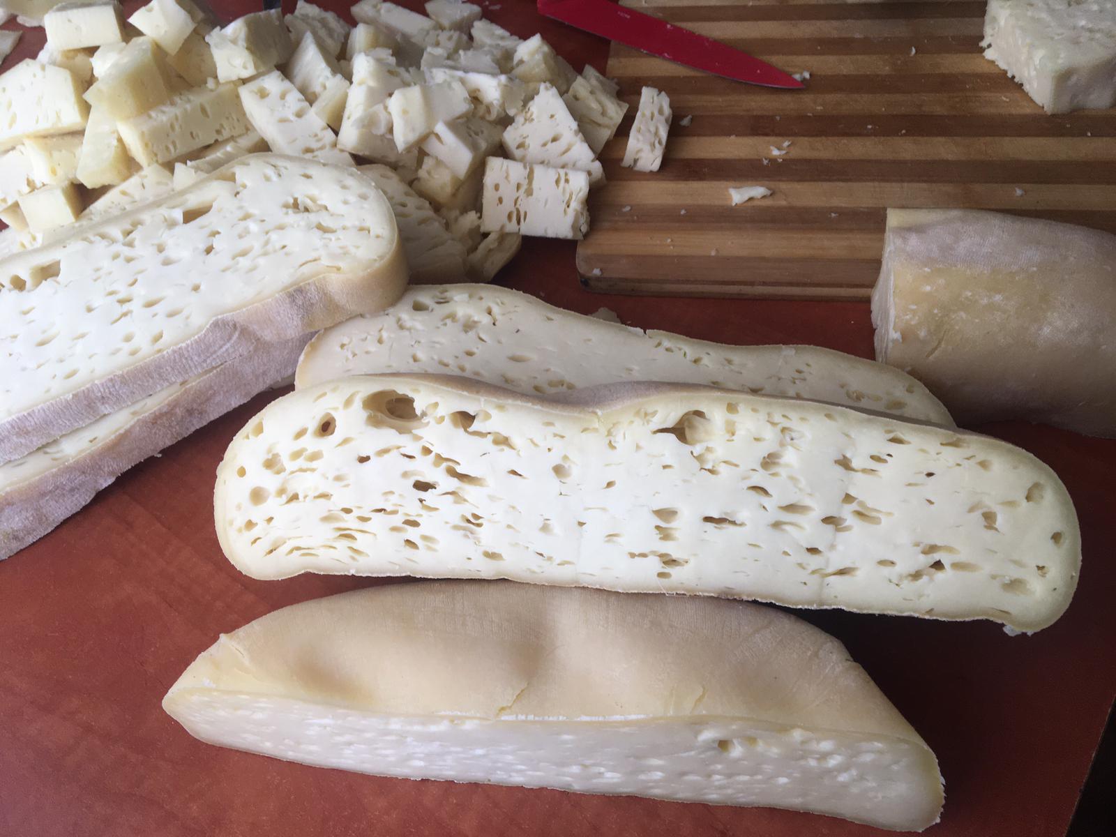Sliced and cubed cheese with a firm, pale yellow texture and small holes, arranged on a wooden cutting board. A red-handled knife is visible in the background.