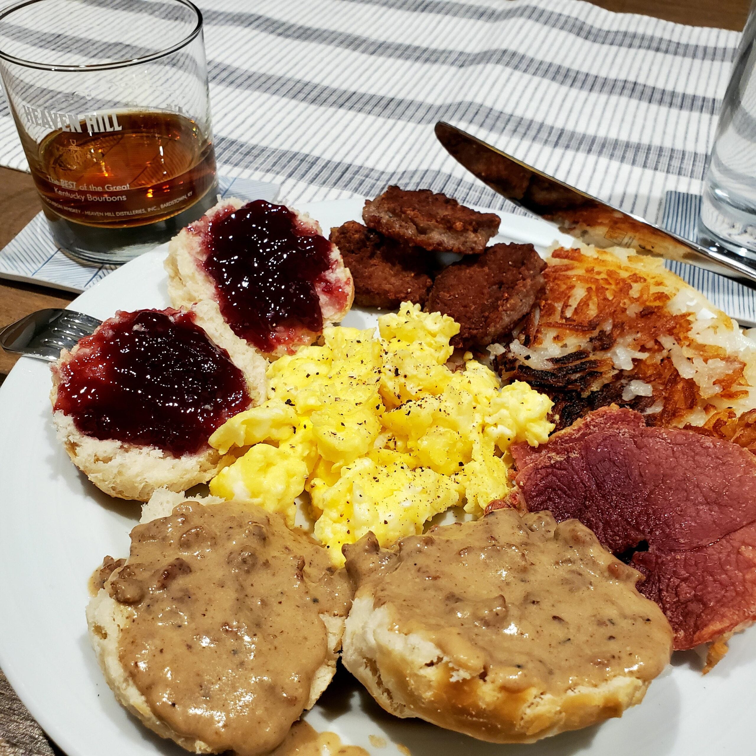 A breakfast plate with scrambled eggs, hash browns, biscuits with gravy, biscuits with jelly, sausage patties, and sliced ham. A glass of whiskey and a glass of water are beside the plate on a striped placemat.