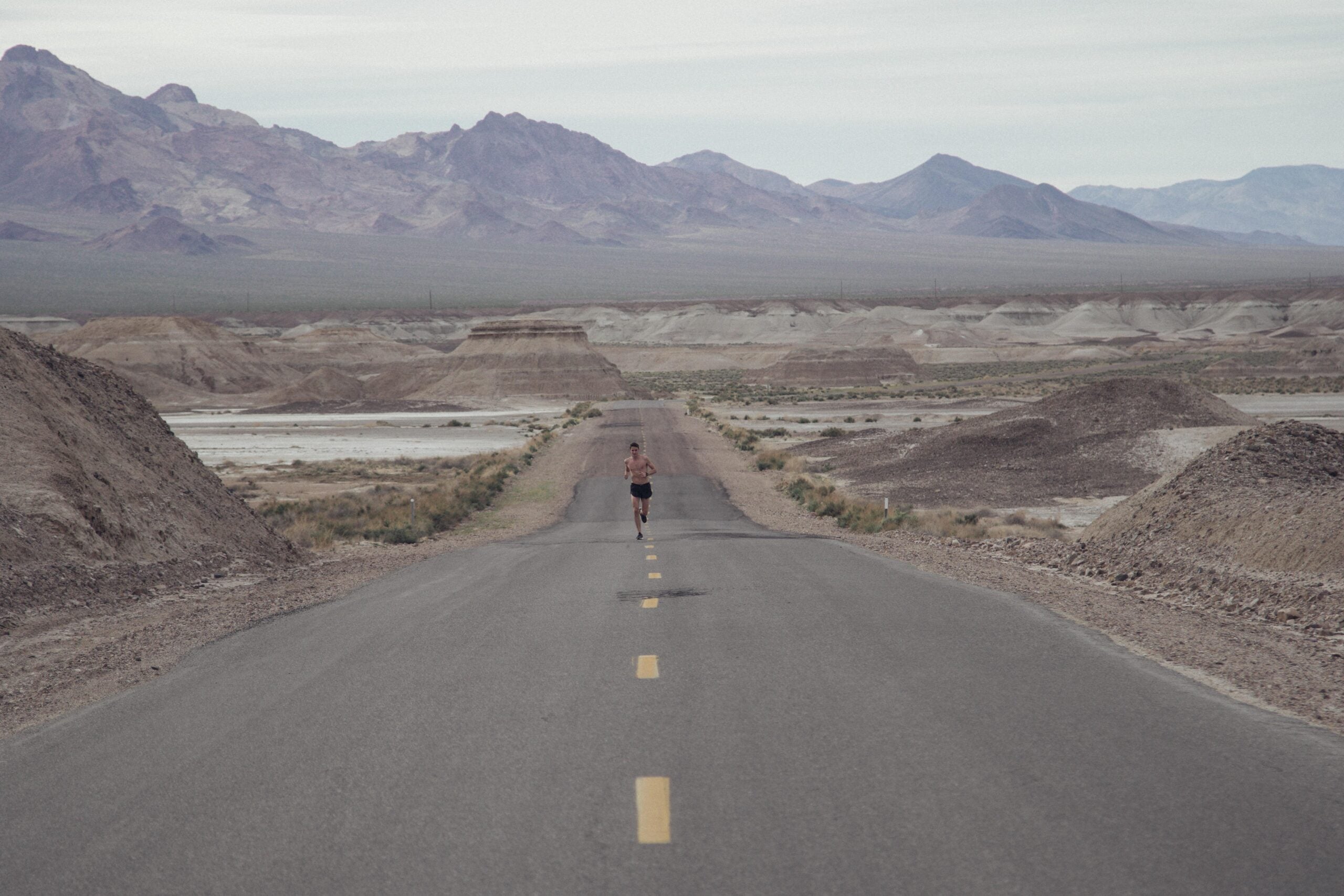 A person runs alone on a long, empty road through a barren desert landscape, with mountains in the background under a cloudy sky.
