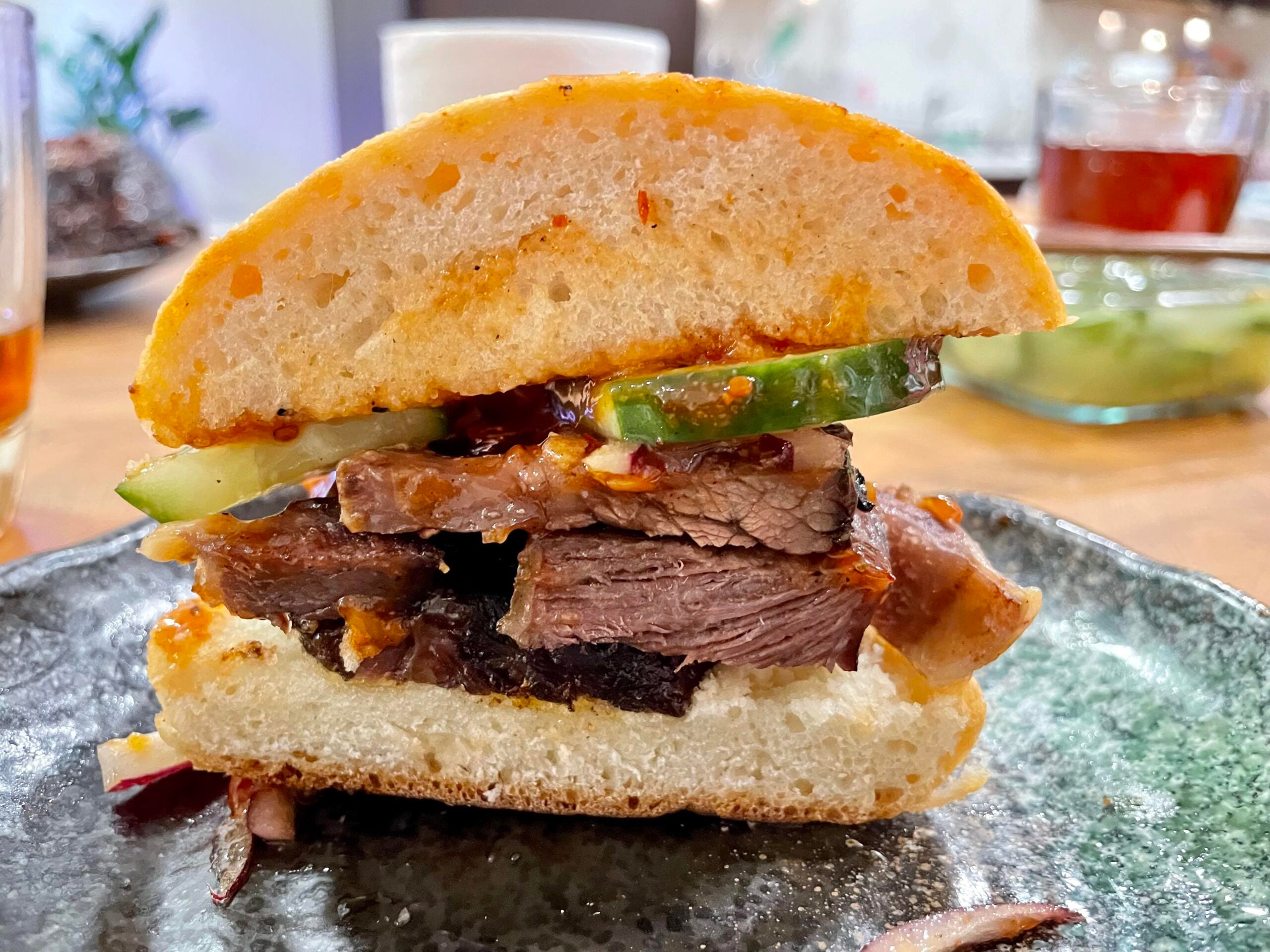 A close-up of a sandwich with thick slices of beef, cucumber, and sauce on a round bun, served on a dark ceramic plate.