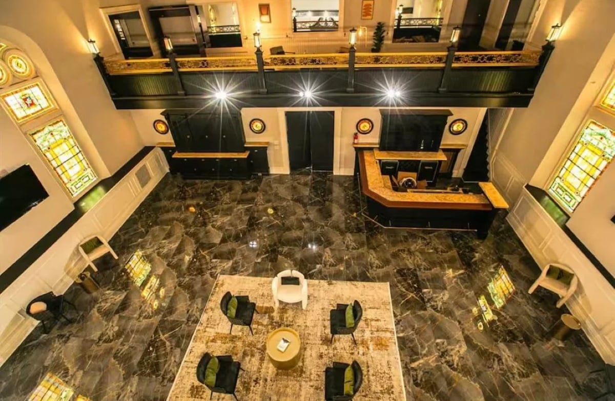A luxurious hotel lobby with marble floors, a central seating area with chairs around a rug, a reception desk, and stained glass windows. The photo is taken from an upper level overlooking the space.