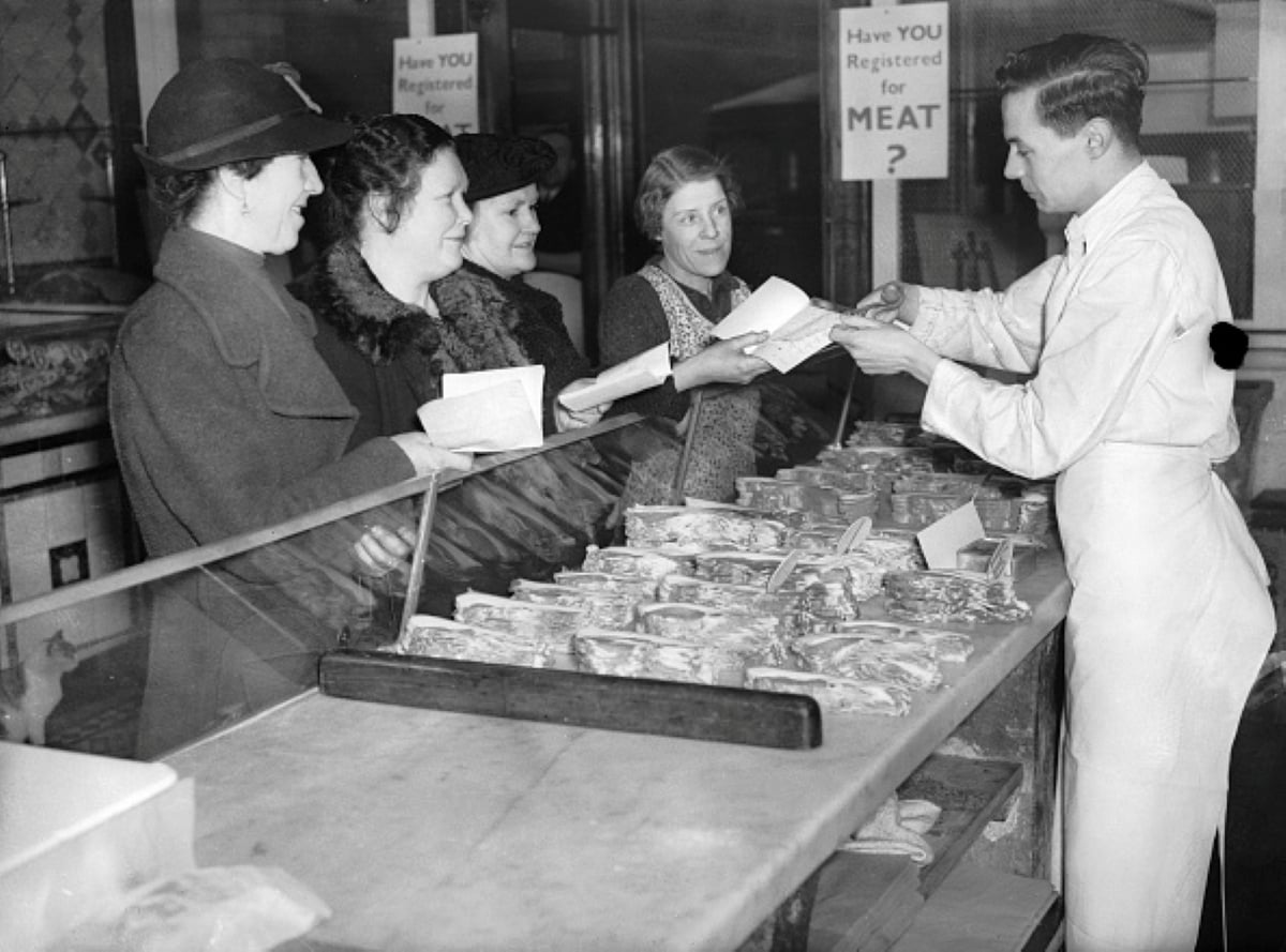 Four women stand in line with ration books at a butcher’s shop counter, receiving packaged meat from a male employee in a white apron. Signs behind them ask, “Have YOU Registered for MEAT?” This scene evokes meals with historical significance, capturing the everyday realities of wartime rationing.