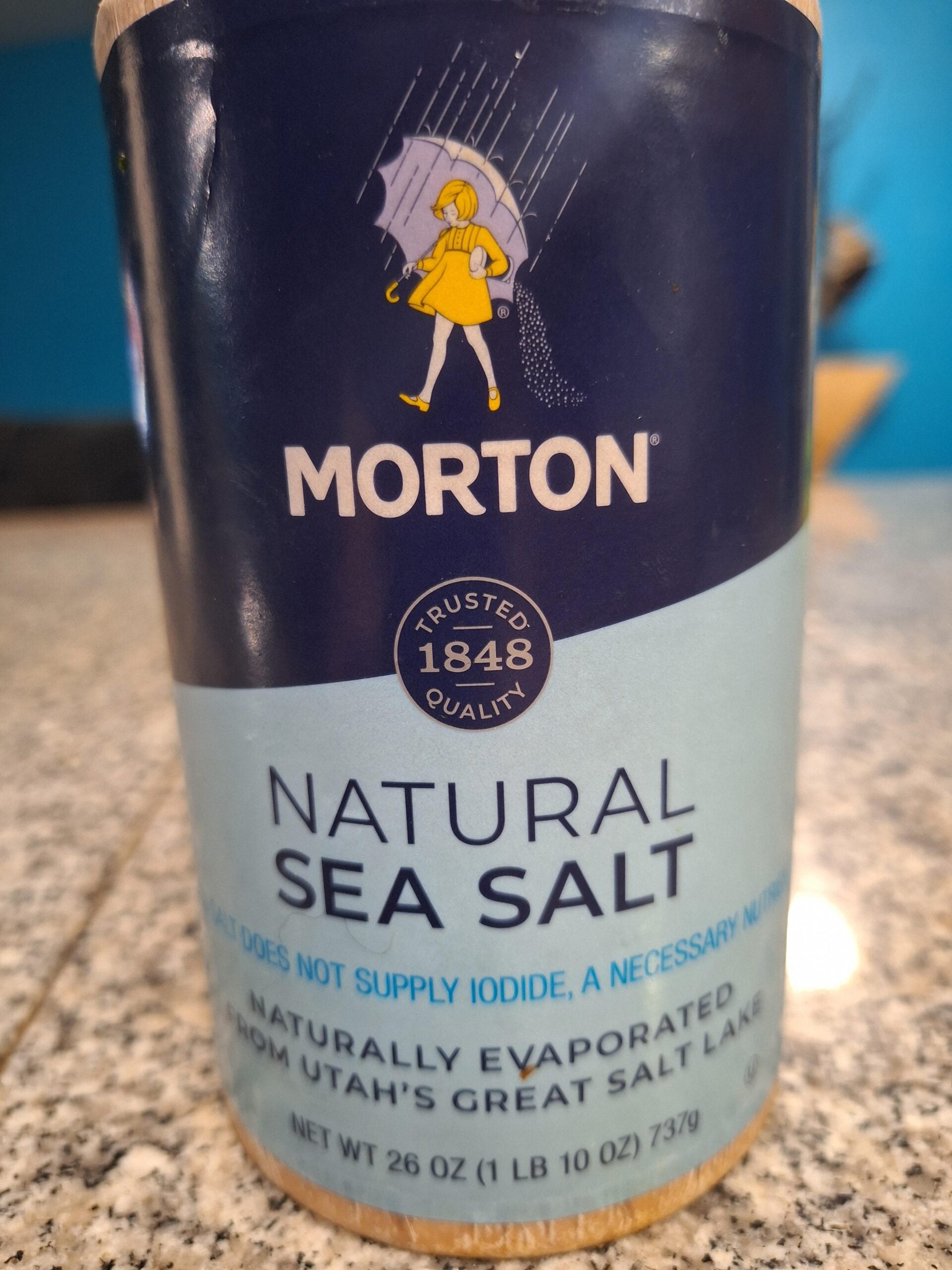 A Morton Natural Sea Salt container sits on a granite counter, featuring the yellow girl with an umbrella logo and blue packaging.