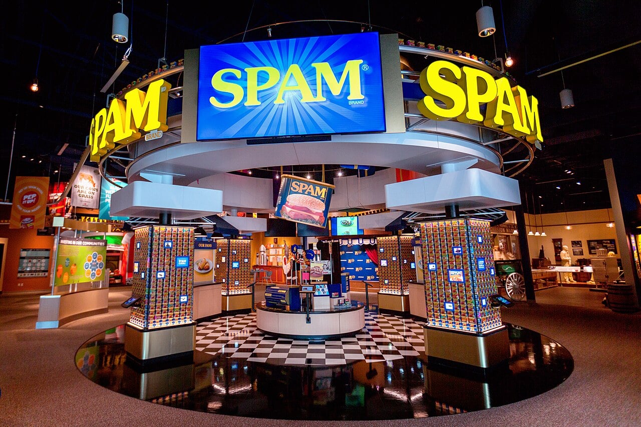 A brightly lit museum exhibit features large yellow “SPAM” signs, blue displays, and shelves filled with SPAM cans, celebrating the iconic canned meat brand in a vibrant, interactive setting.