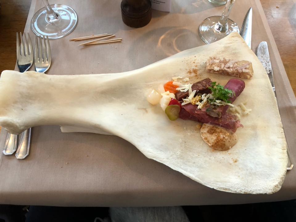 A gourmet appetizer with meats and vegetables is artfully arranged on a large, flat bone, set on a table with cutlery, wine glasses, and toothpicks nearby.