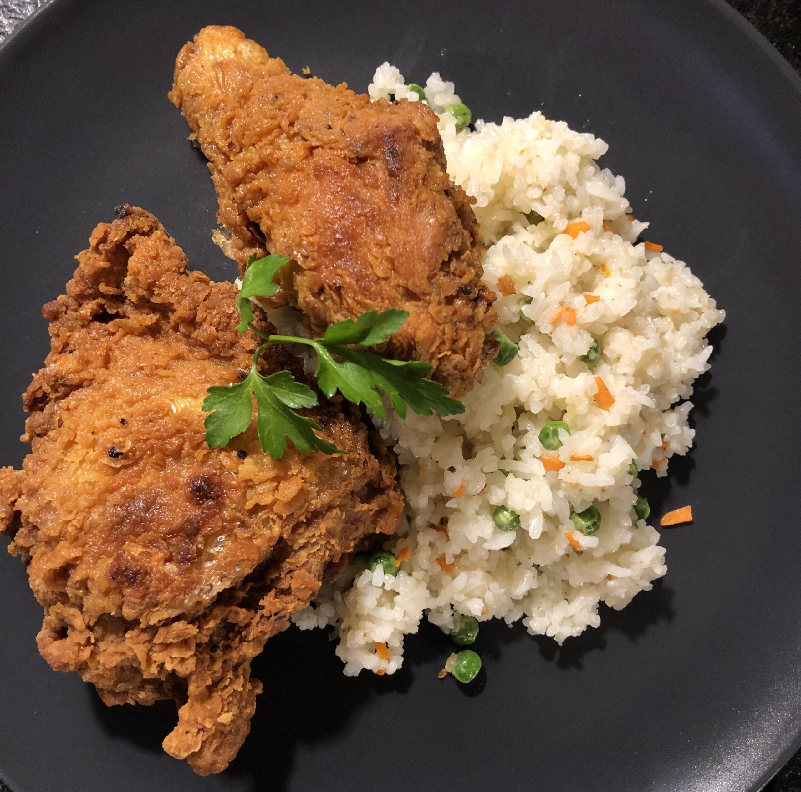 Two pieces of crispy fried chicken served with white rice mixed with peas and diced carrots, garnished with a sprig of parsley, on a black plate.