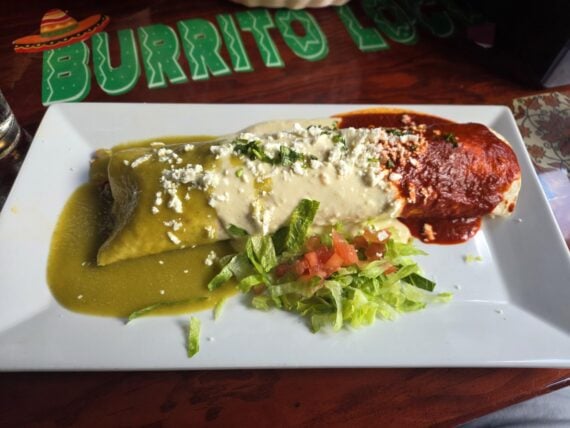 A burrito topped with green and red sauces, white crema, and crumbled cheese, served with shredded lettuce and pico de gallo on a rectangular white plate.