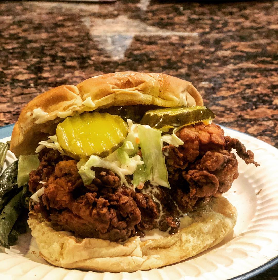 A fried chicken sandwich with pickles, lettuce, and sauce on a bun, served on a white plate with a side of green beans on a marbled countertop.