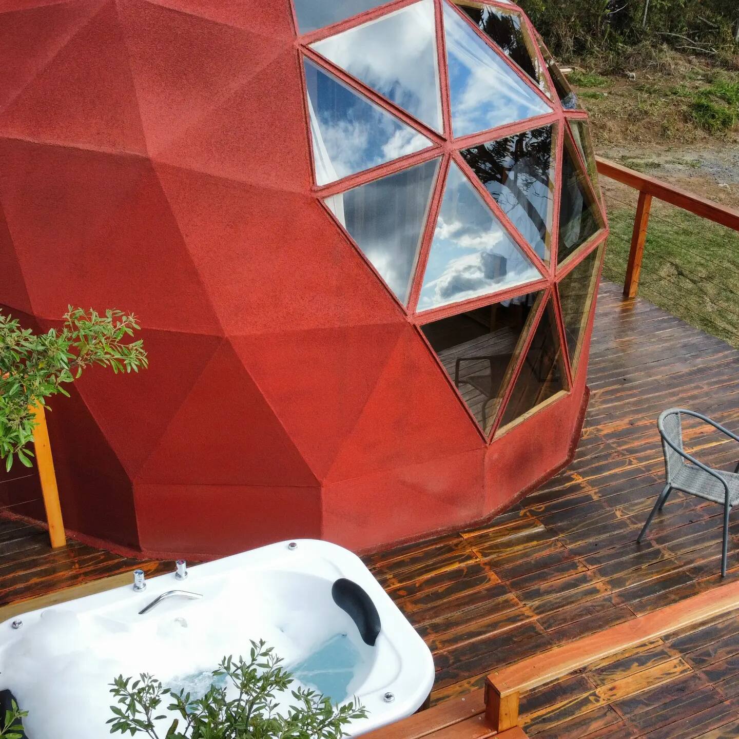 A red geodesic dome with triangular windows sits on a wooden deck with a white outdoor jacuzzi and a metal chair. Trees and grass are visible around the structure.