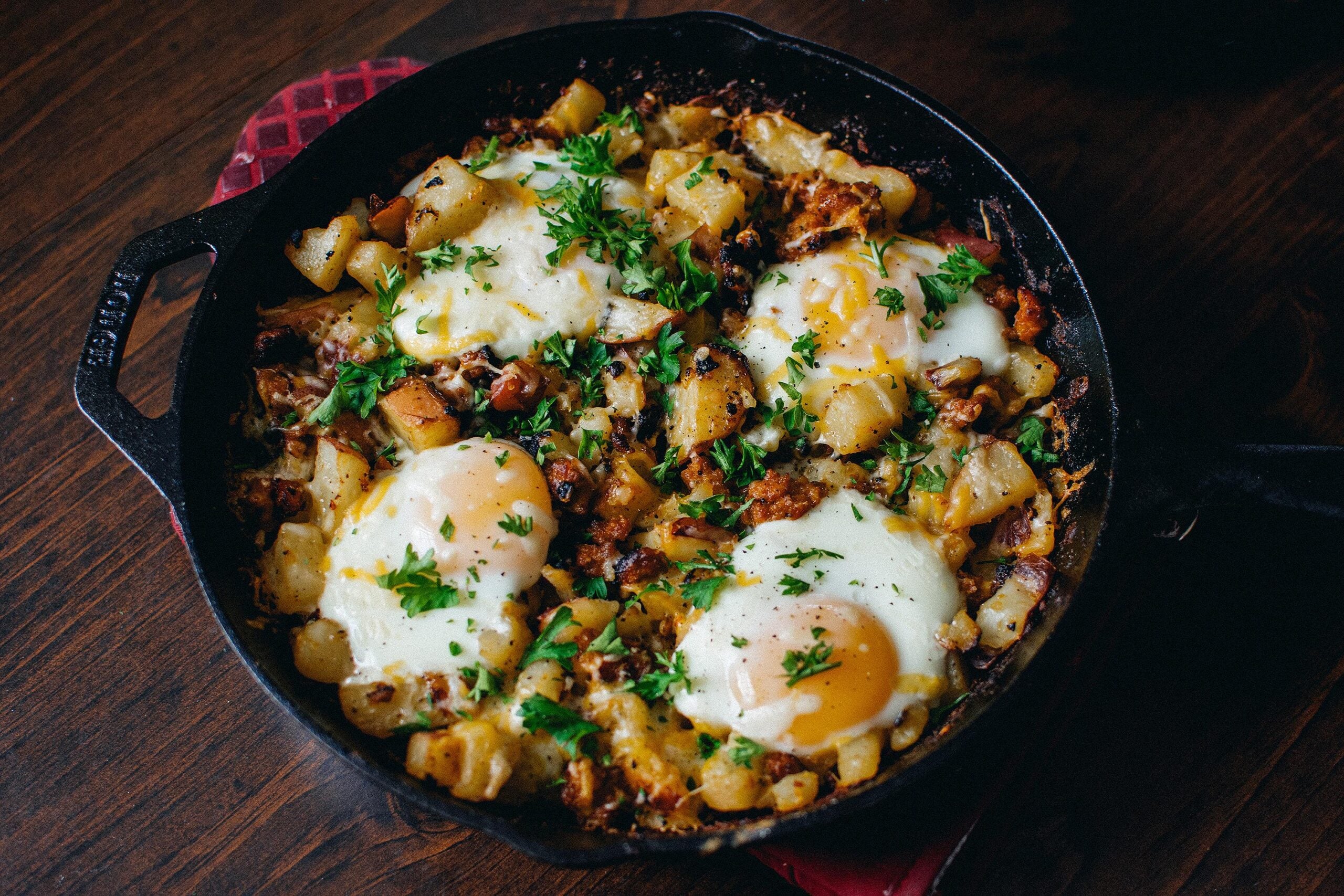 A cast iron skillet filled with hash made of potatoes, onions, and three sunny-side-up eggs, garnished with chopped parsley, sits on a wooden table.