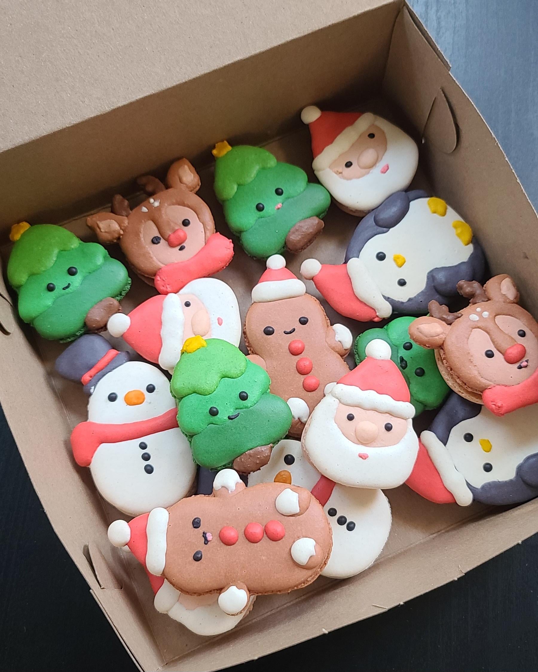 A box of colorful macarons, made as delightful homemade Christmas treats and decorated as Christmas characters—including Santa, reindeer, snowmen, Christmas trees, and gingerbread men—arranged neatly inside a brown cardboard box.