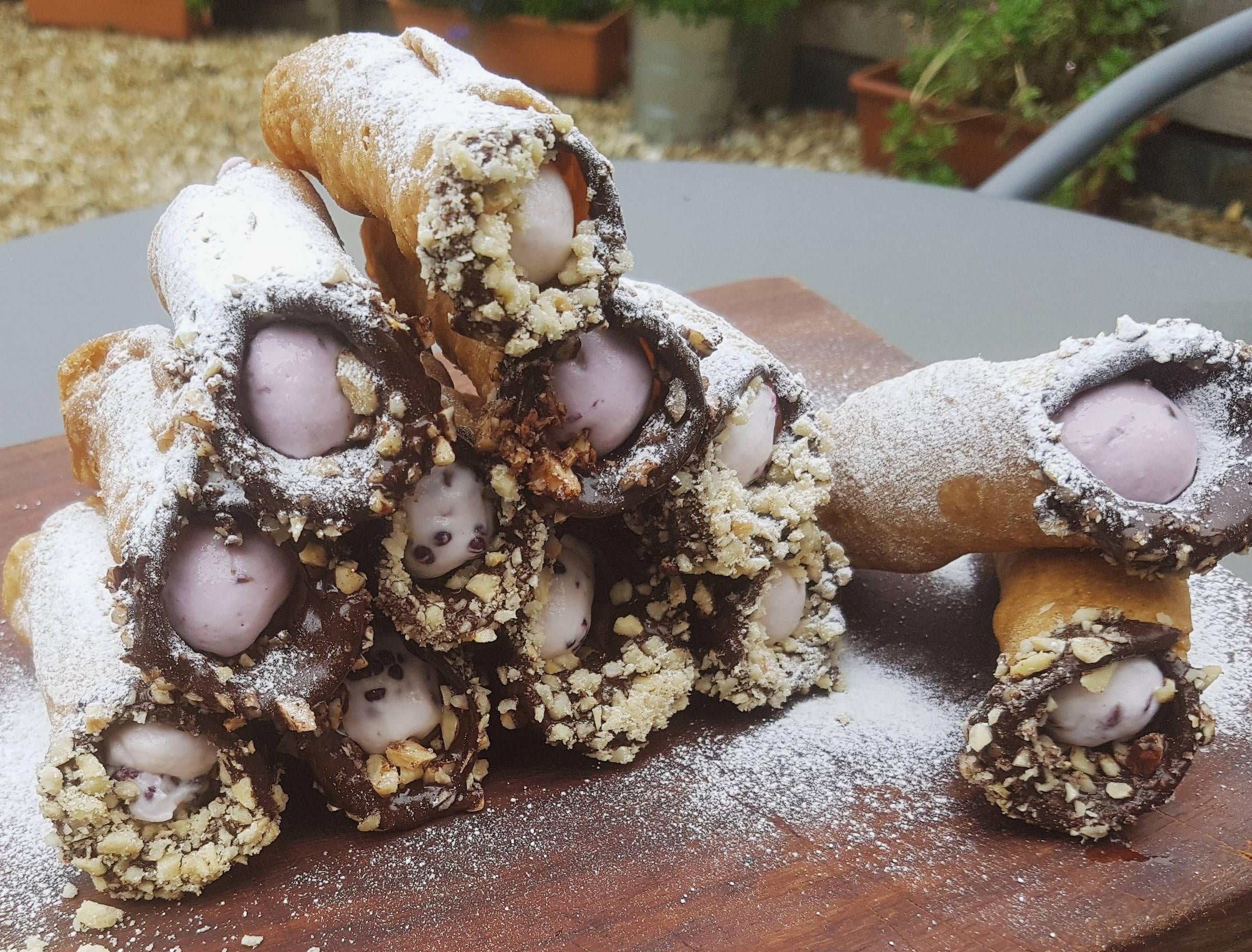 A stack of homemade cannolis filled with purple and white cream, dusted with powdered sugar and rolled in chopped nuts, displayed on a wooden board outdoors.