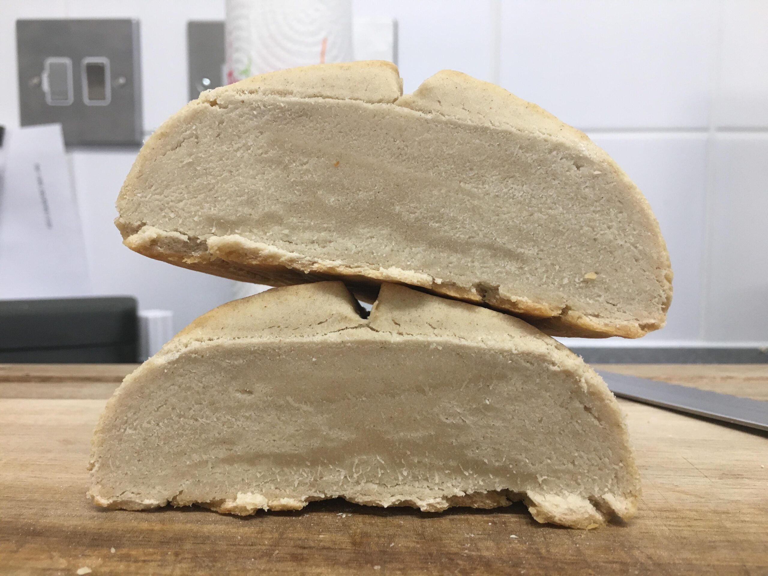 Two thick, pale, dense halves of a loaf of bread are stacked on a wooden surface, revealing a compact, underbaked interior with a hard crust. The background shows a kitchen counter, switches, and a paper towel roll.