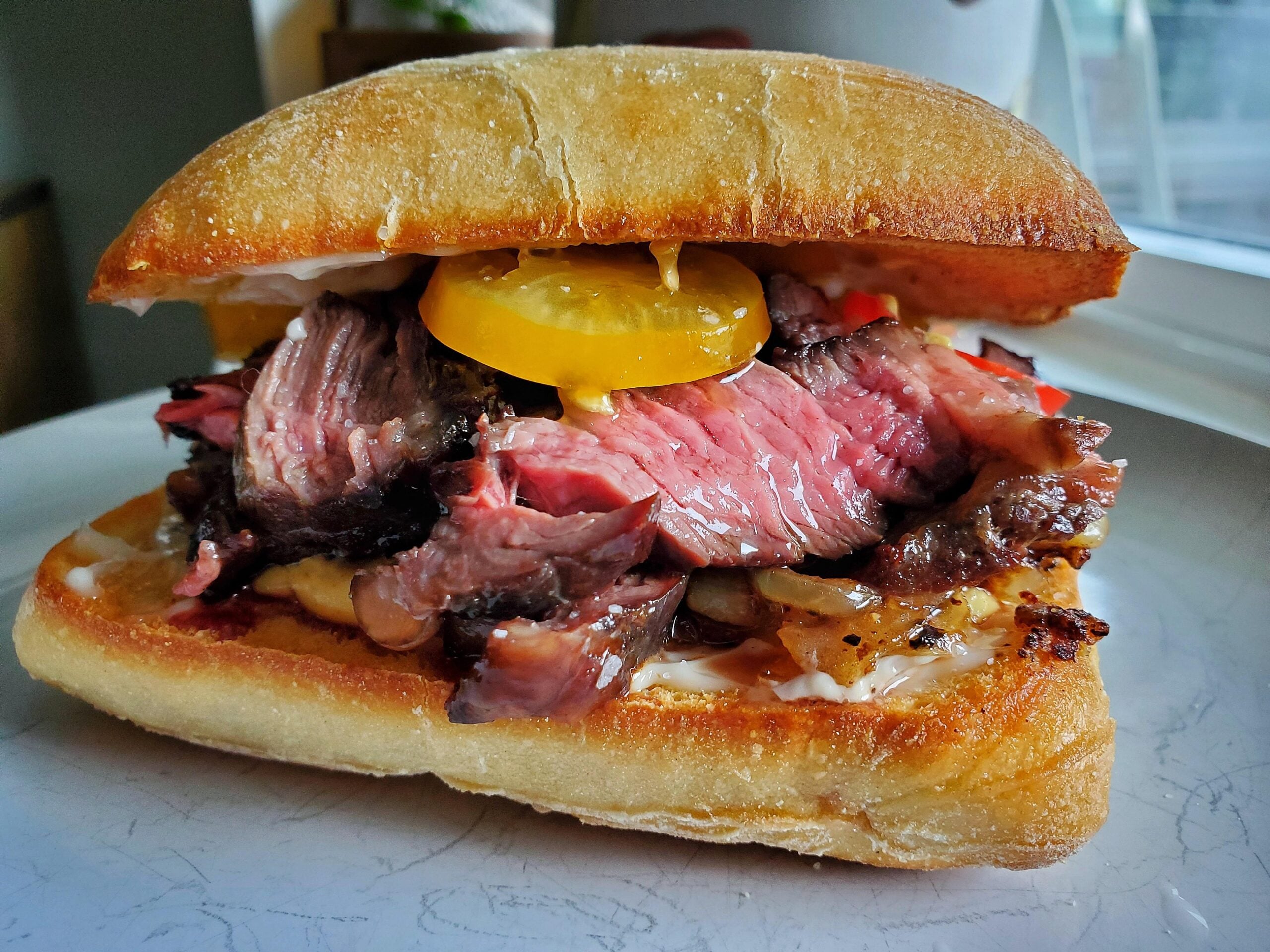 A close-up of a steak sandwich on ciabatta bread, filled with sliced medium-rare steak, yellow tomato, onions, and mayonnaise, served on a white plate.