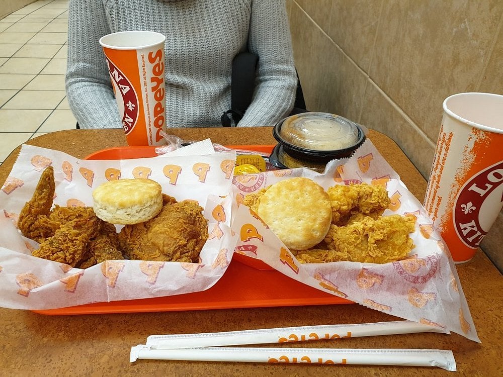Two trays of fried chicken, biscuits, and mashed potatoes with gravy on a restaurant table. A person sits behind the food, partly visible, wearing a gray sweater. Two soft drink cups and straws are also on the table.