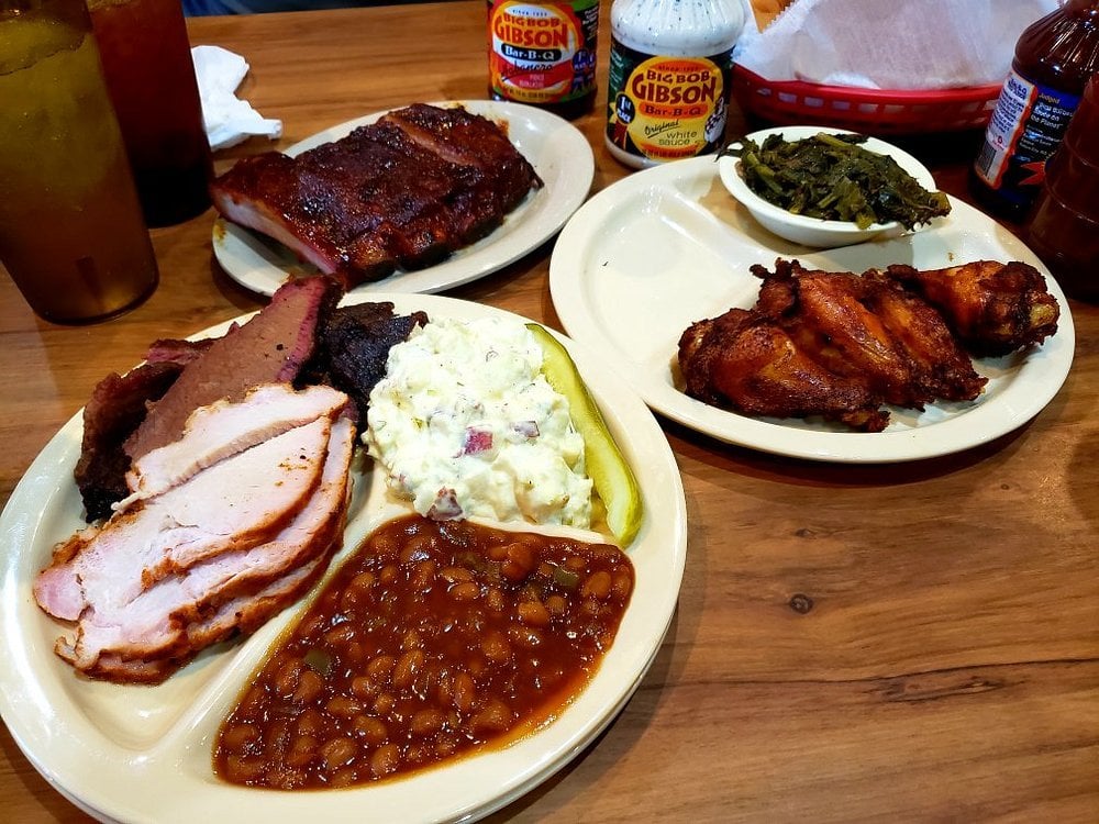 Barbecue plate with sliced turkey, brisket, baked beans, potato salad, pickle, ribs, chicken wings, collard greens, and sauces on a wooden table.