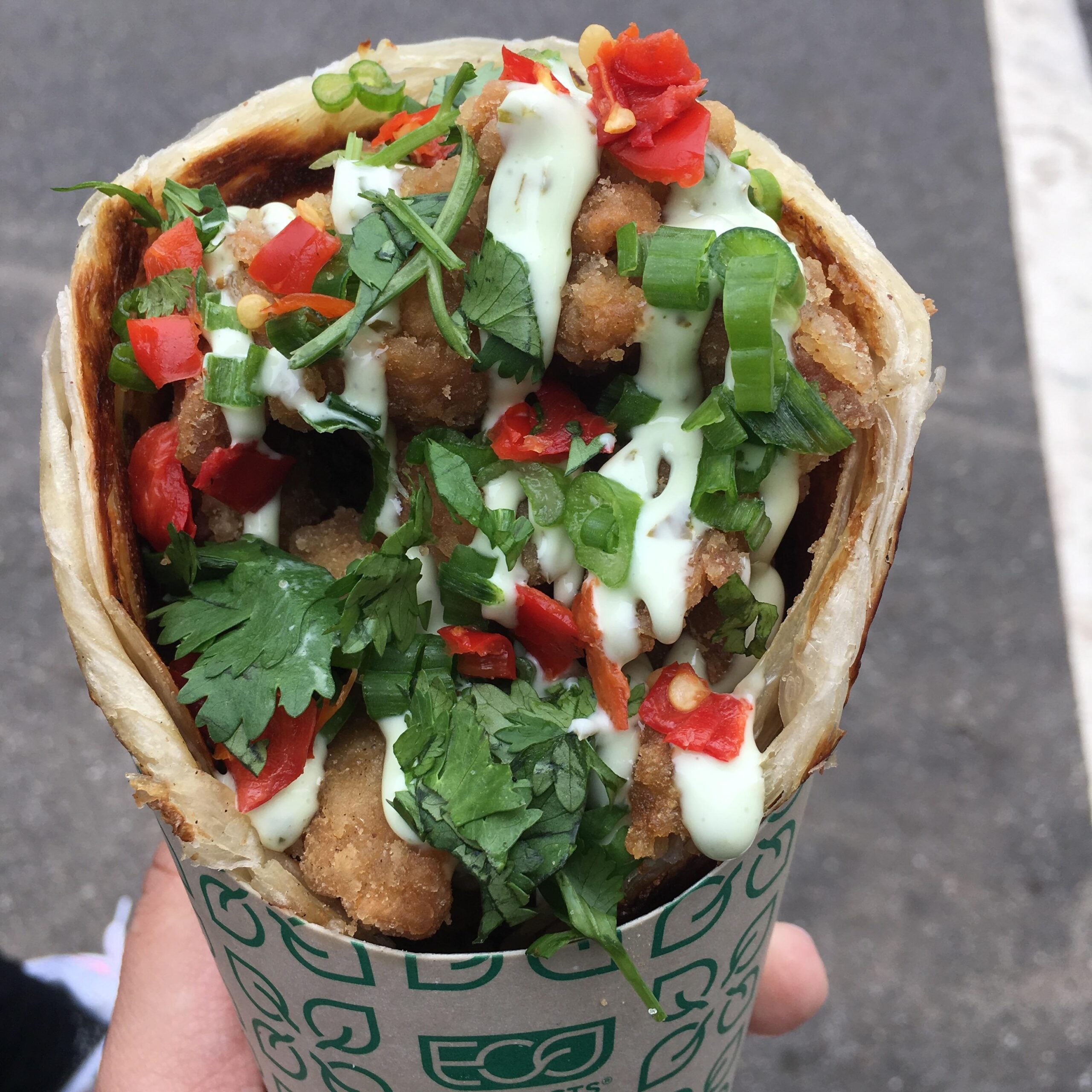 A close-up of a hand holding a wrap filled with fried chicken, chopped red peppers, green onions, cilantro, and a drizzle of creamy white sauce.